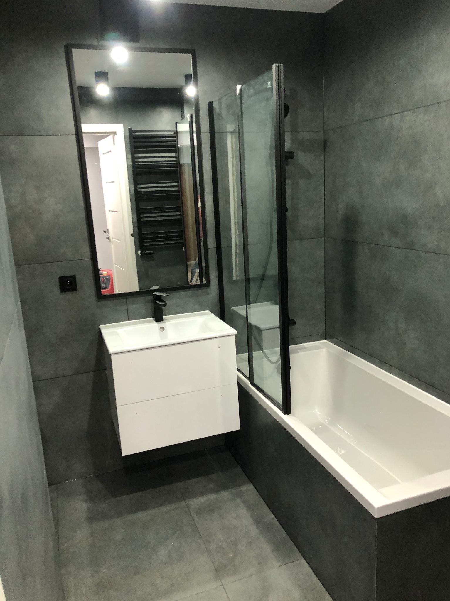 Nowoczesna łazienka w szarościach: biała umywalka z szafką, czarne detale, przeszklona kabina nad wanną, duże szare płytki na ścianach i podłodze.