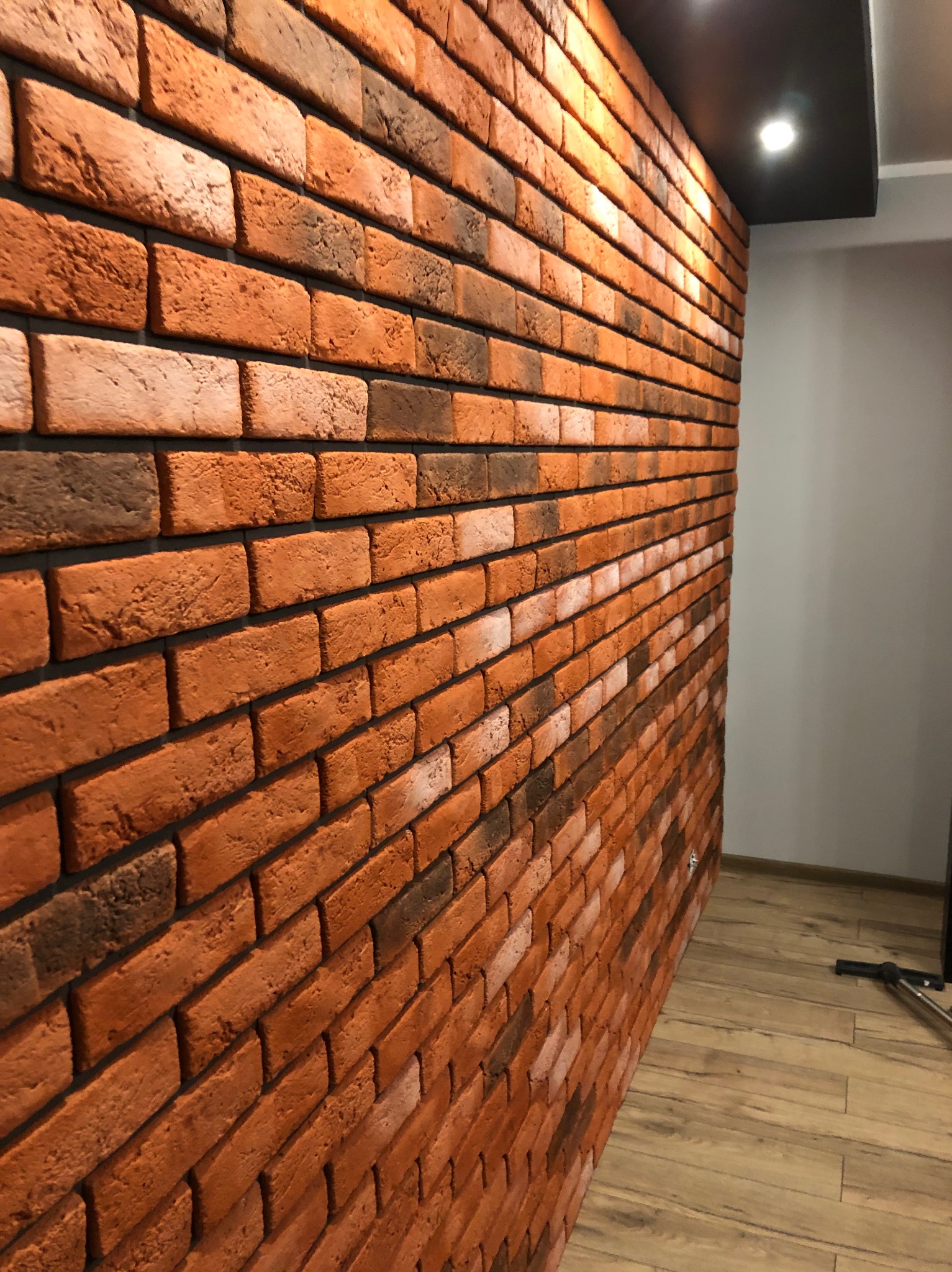 Dekoracyjna ściana z cegły w odcieniach brązu i pomarańczy, ułożona w regularny wzór, oświetlona punktowym światłem w pomieszczeniu z drewnianą podłogą.