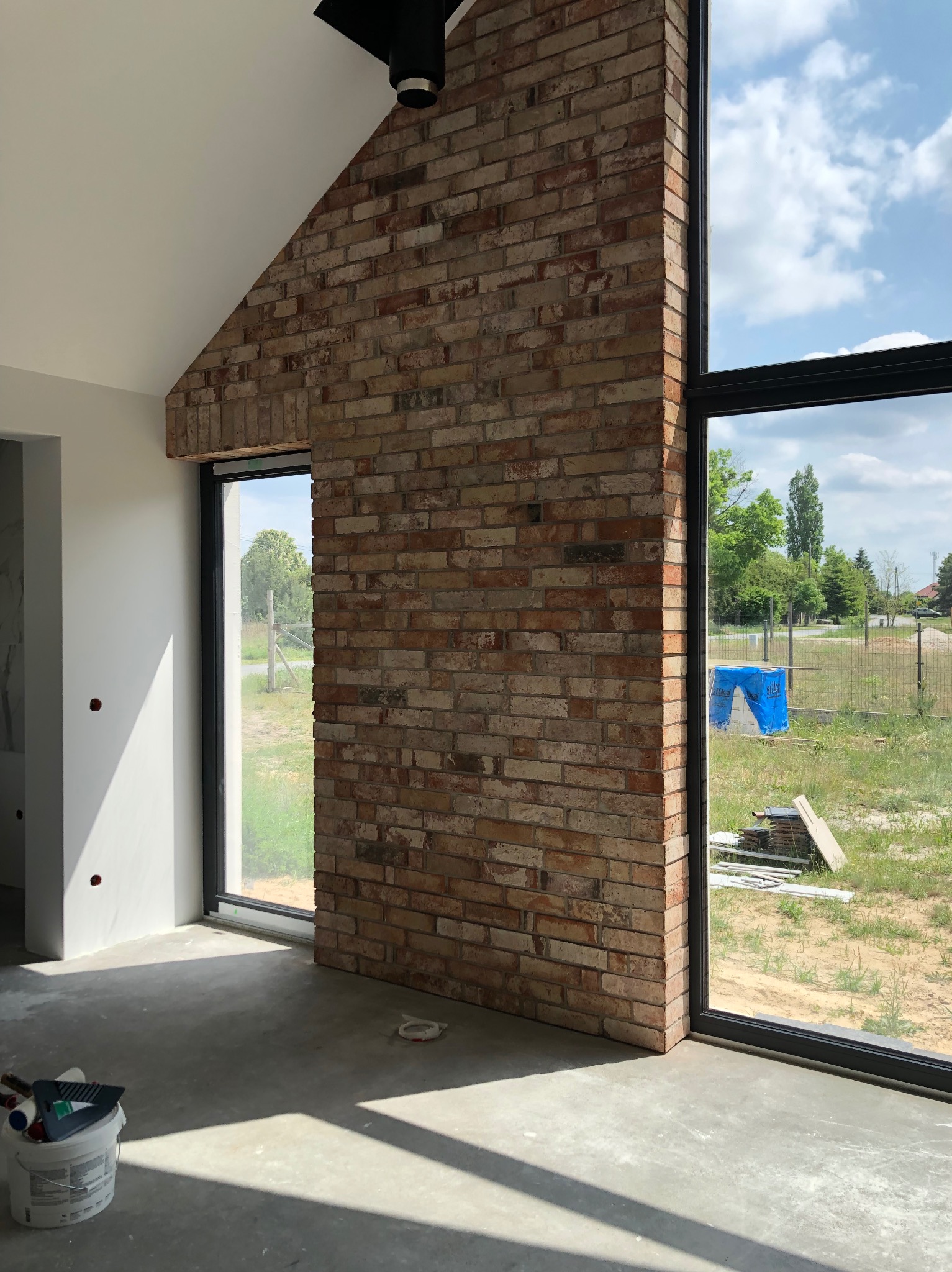 Wnętrze z ceglaną ścianą, dużymi oknami i surowym betonem. Widoczne narzędzia malarskie. Cegła tworzy trójkątny kształt, wpisując się w architekturę pomieszczenia.