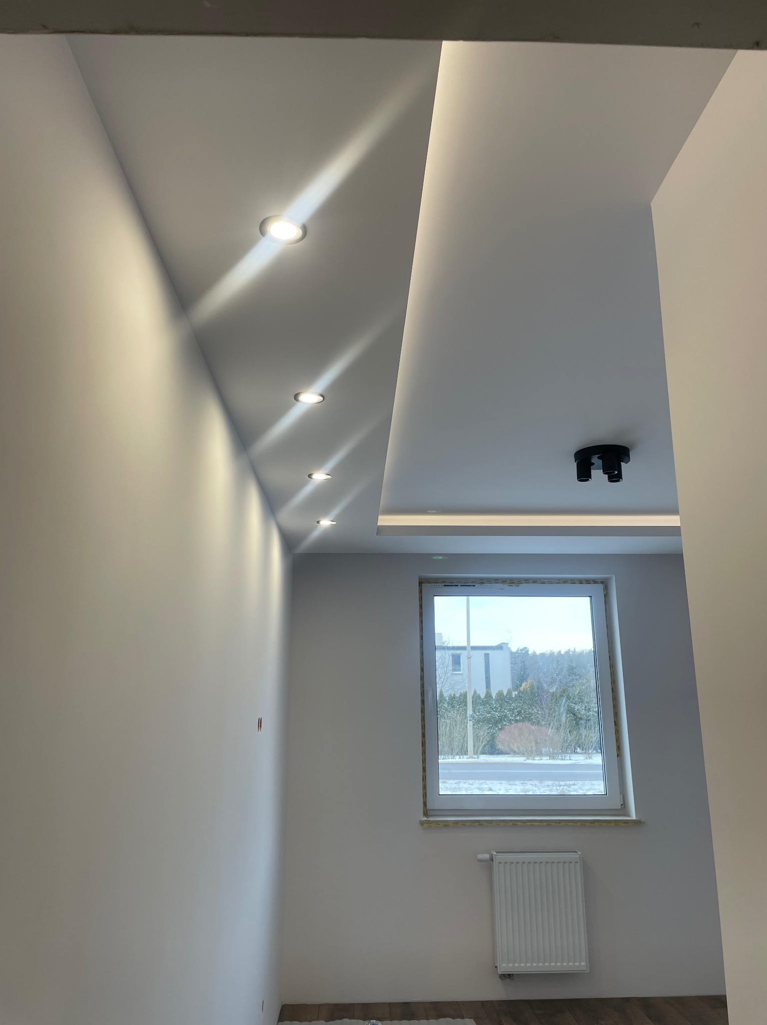 Nowoczesne wnętrze: białe ściany, sufit podwieszany z oświetleniem punktowym, okno z widokiem na zewnątrz, grzejnik. Minimalizm i jasne barwy.