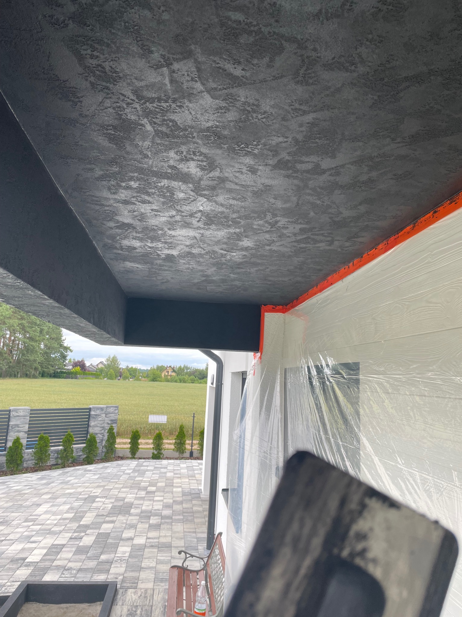 Wykończony sufit z efektem dekoracyjnym w odcieniach szarości, widoczny fragment elewacji zabezpieczony folią malarską i narzędzie w dolnym rogu.