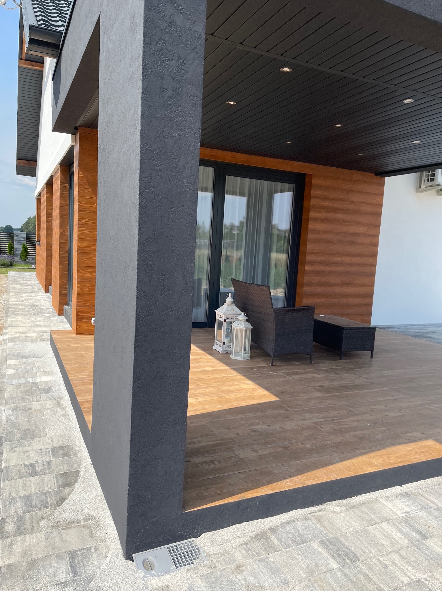 Nowoczesny taras z drewnianą podłogą i elewacją, szary filar z fakturą, meble ogrodowe i lampiony. Widok na zadbany ogród w tle.