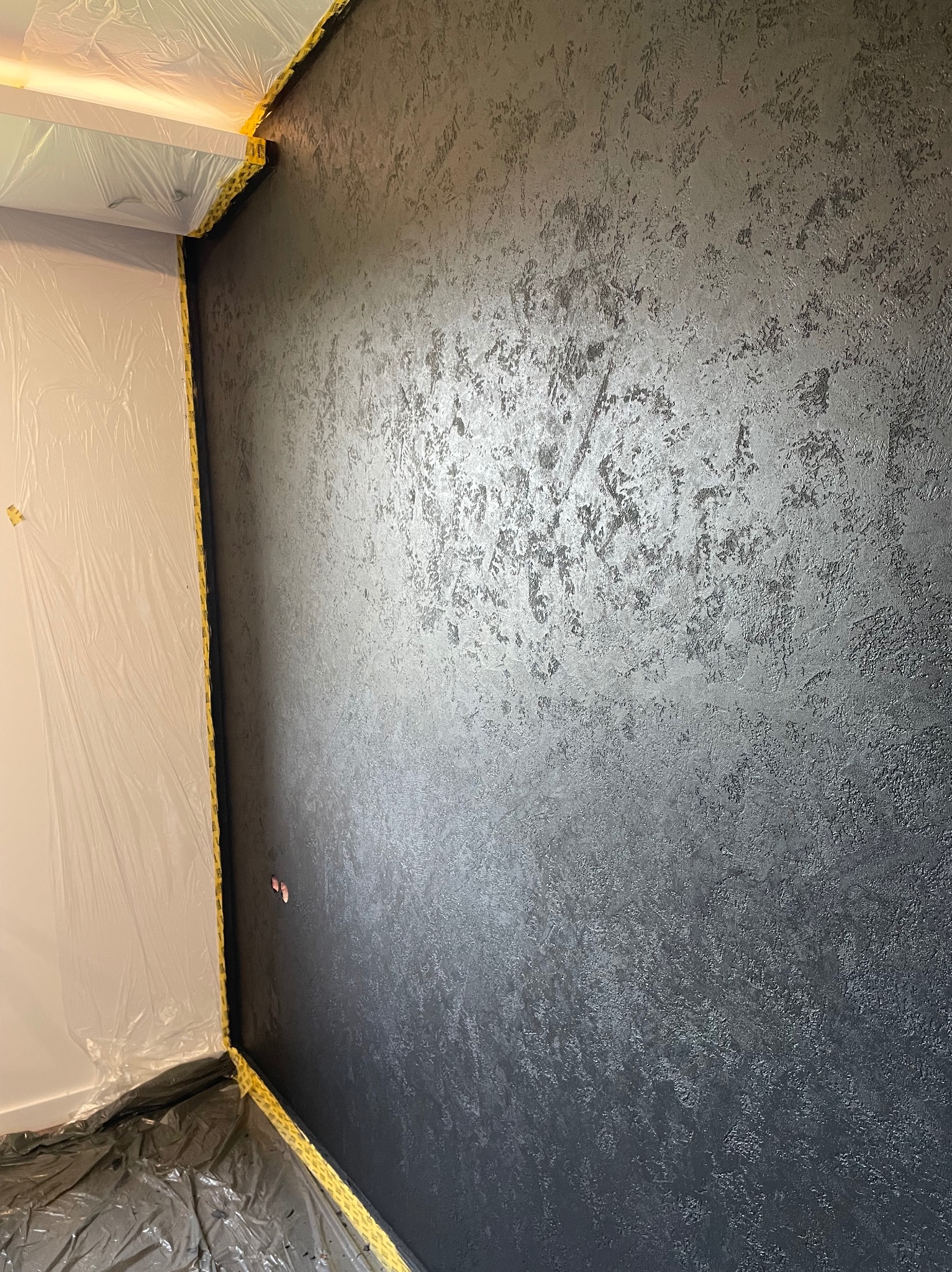 Ściana z dekoracyjną, ciemną fakturą, zabezpieczona folią i taśmą malarską. Widoczne detale wykończenia i przygotowanie do dalszych prac.