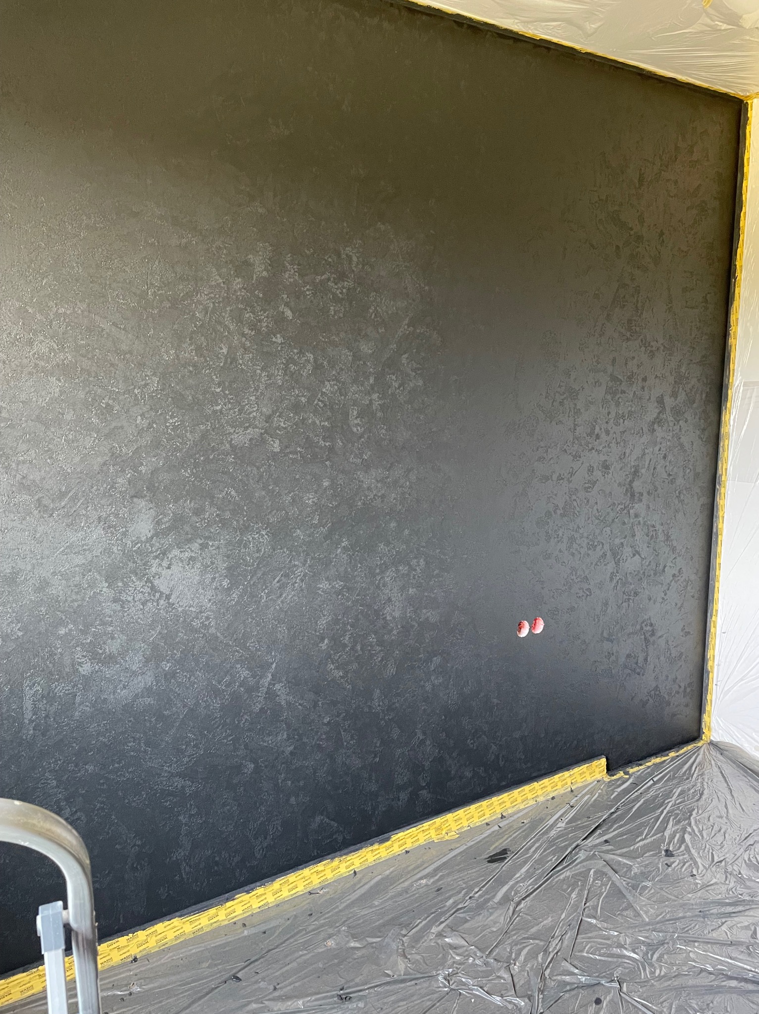 Ściana w pokoju po nałożeniu dekoracyjnej, ciemnej gładzi w stylu weneckim. Zabezpieczone folią podłoga i ściany. Widoczne gniazdka elektryczne i fragment drabiny.