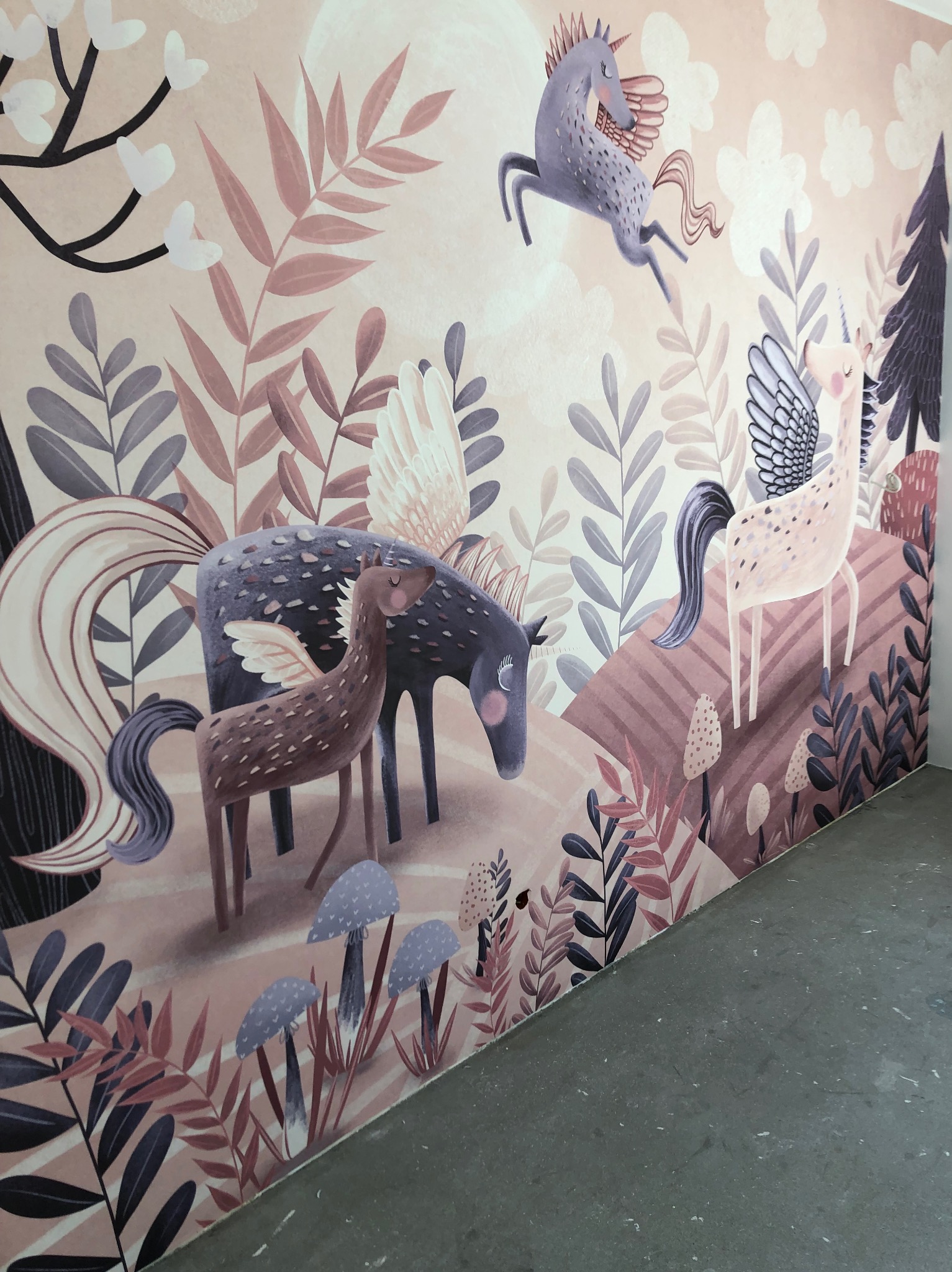 Ściana z dziecięcą tapetą przedstawiającą jednorożce w leśnej scenerii, utrzymana w pastelowych odcieniach różu, fioletu i brązu. Widoczne liście, grzyby i drzewa.