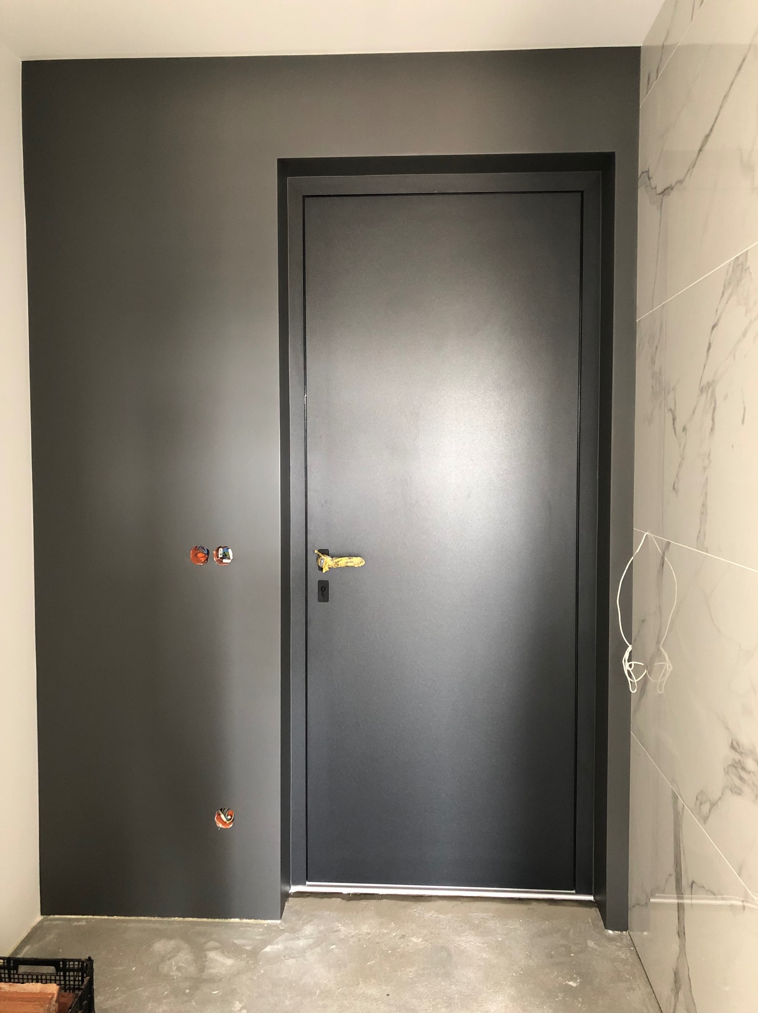 Ciemne drzwi z złotą klamką w nowym wnętrzu. Ściana pomalowana na szaro, obok marmurowe płytki. Widoczne gniazdka elektryczne przed wykończeniem.