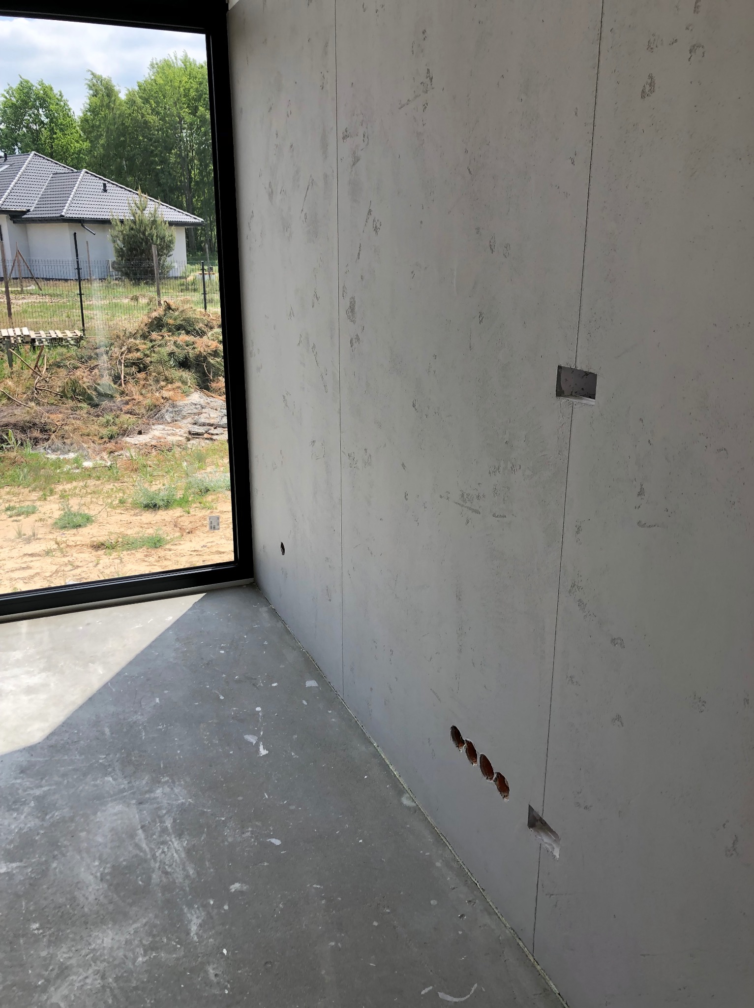 Szara ściana z wywierconymi otworami na gniazdka elektryczne, surowy beton na podłodze, widok na zewnątrz przez duże okno. Wnętrze w trakcie wykańczania.