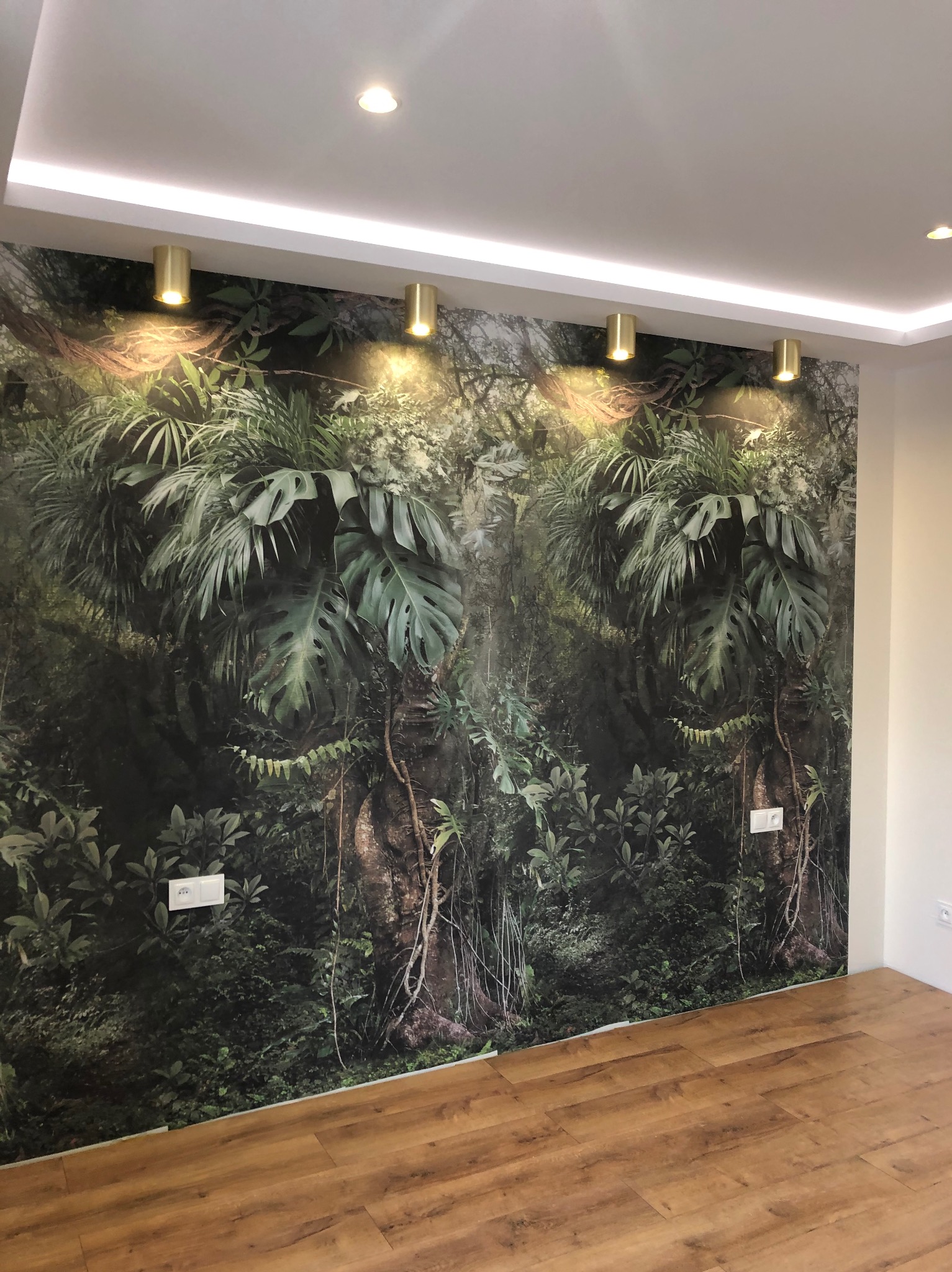 Ściana z fototapetą imitującą dżunglę, oświetlona złotymi lampami tubowymi. Widoczna drewniana podłoga i biały sufit z listwą LED. Nowoczesne wykończenie wnętrza.