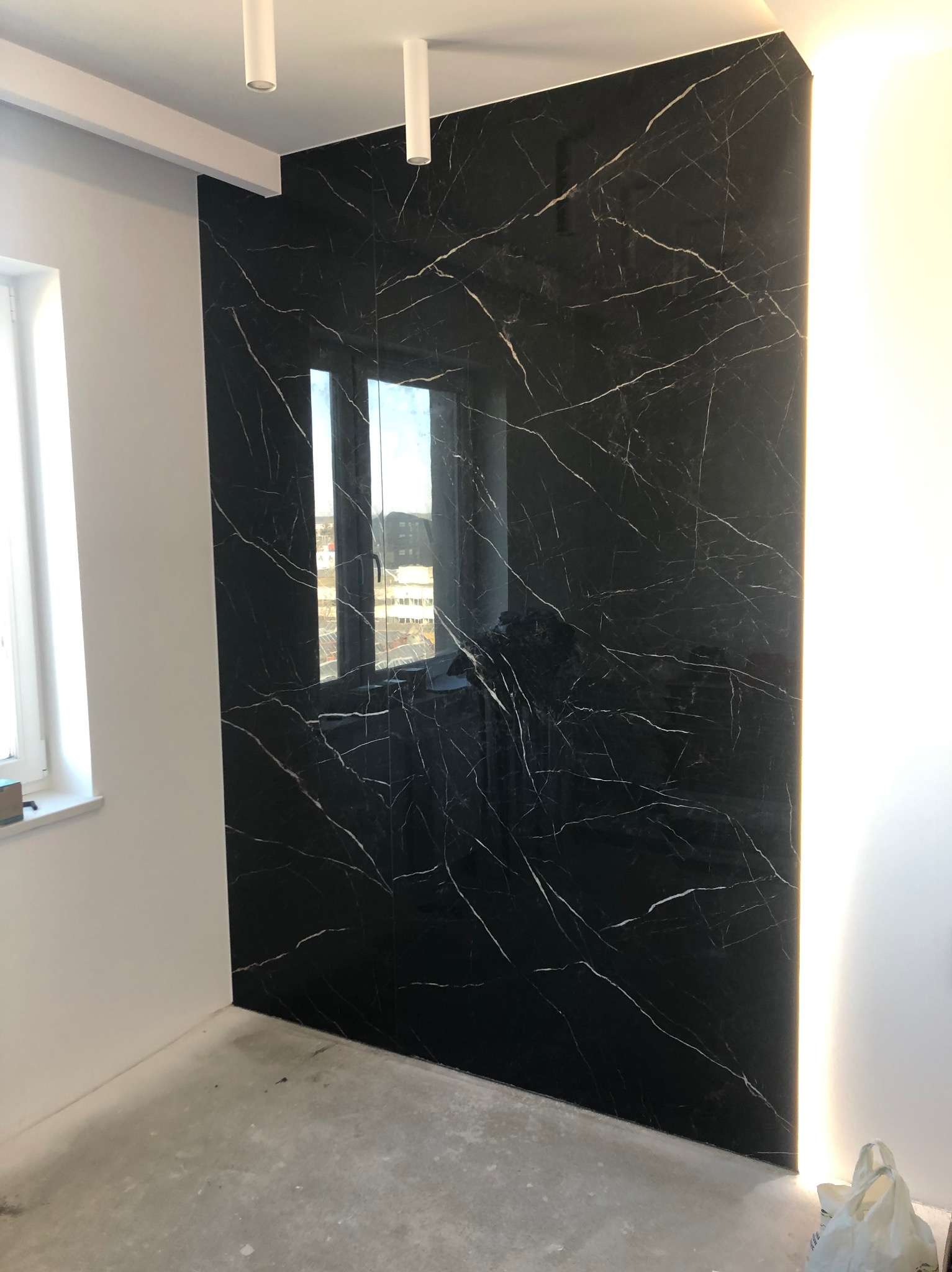 Ściana wykończona czarnymi płytami imitującymi marmur z białymi żyłami, podświetlona LED w Golub-Dobrzyń. Widoczne okno i sufit z oświetleniem.