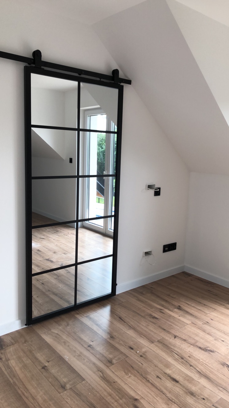 Przesuwne drzwi loftowe z czarną ramą i szprosami, zamontowane na szynie w jasnym pokoju na poddaszu. Widoczne gniazdka elektryczne na ścianie i drewniana podłoga.
