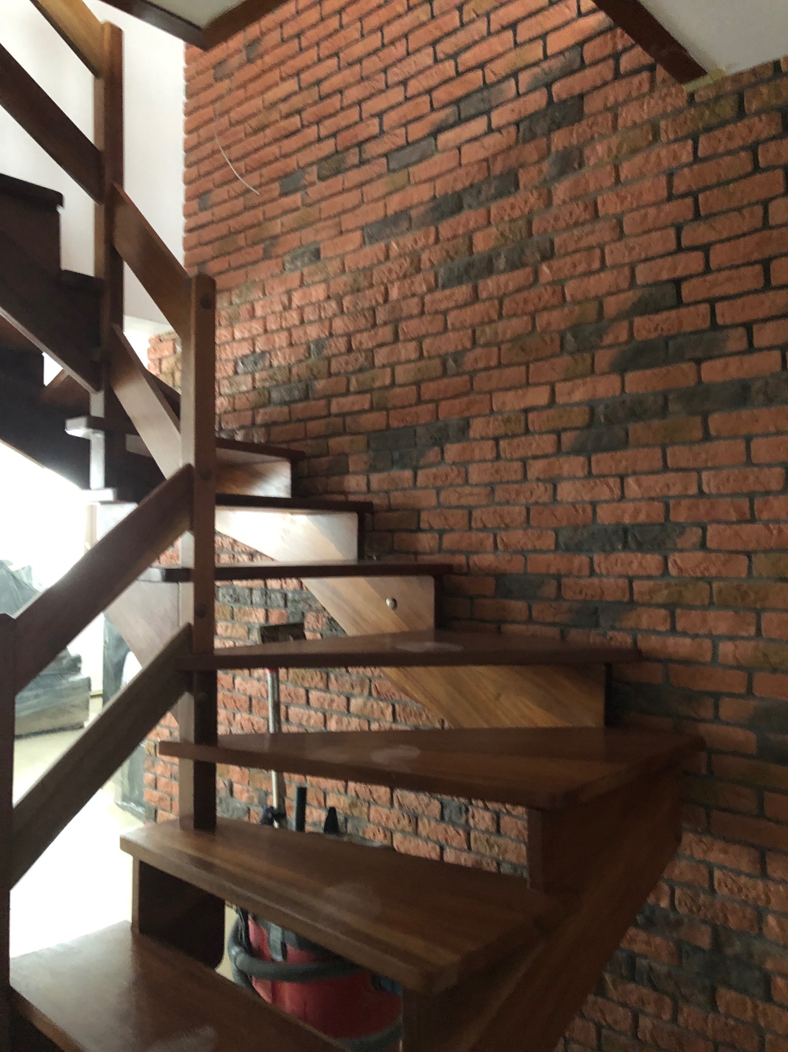 Drewniane schody z balustradą na tle ściany z cegły klinkierowej w odcieniach czerwieni i szarości. Widoczne detale wykończenia i naturalne światło.