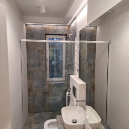 LGK Glazura - Nowoczesna łazienka z kabiną prysznicową typu walk-in, umywalką nablatową i płytkami w odcieniach szarości i brązu, oświetlona listwą LED pod sufitem.