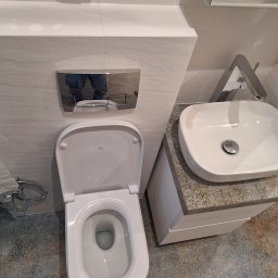 LGK Glazura - Nowoczesna łazienka z białą ceramiką: wisząca miska WC Roca, umywalka nablatowa na szafce z wzorzystym blatem i chromowana bateria, wykończona płytkami w odcieniach szarości.