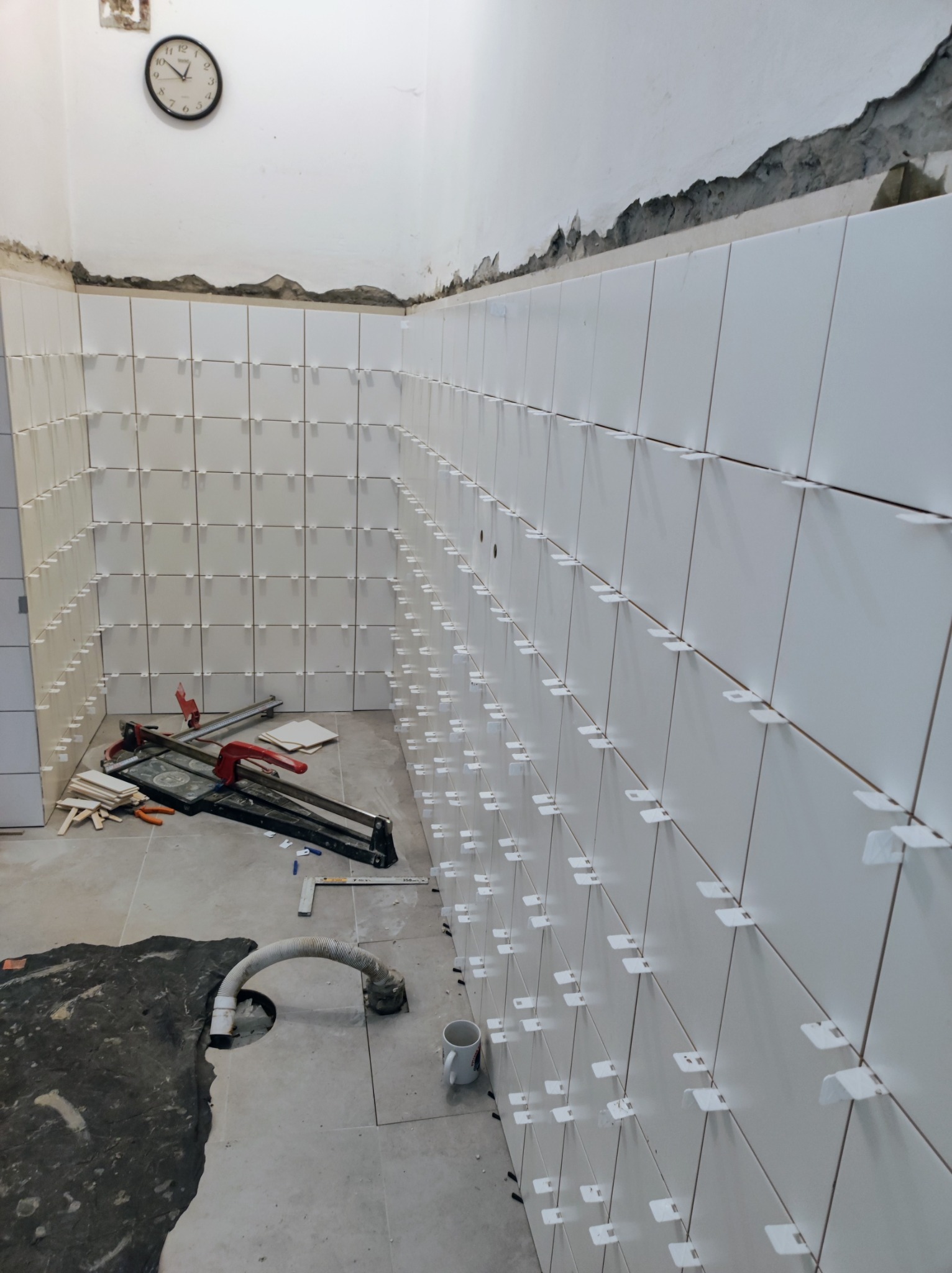 Białe płytki ścienne w trakcie układania w łazience, z widocznymi krzyżykami dystansowymi, narzędziem do cięcia płytek i fragmentem nieukończonej podłogi z rurą odpływową.