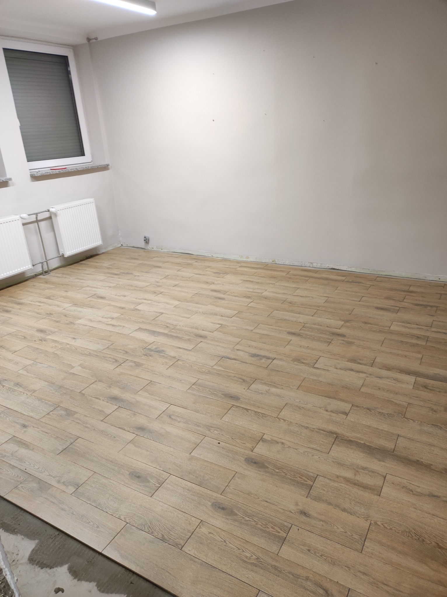 Ułożona podłoga z płytek imitujących drewno w jasnym odcieniu, widoczne grzejniki pod oknem i surowe ściany w pustym pomieszczeniu.