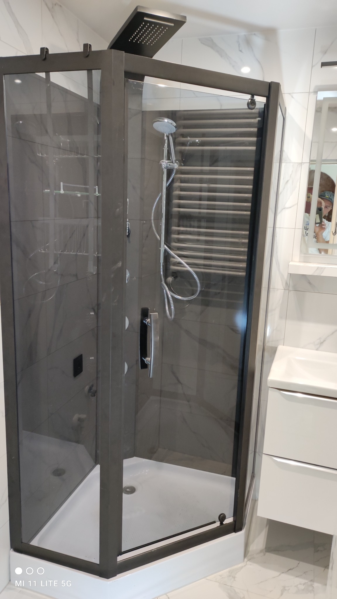 Nowoczesna kabina prysznicowa z ciemną ramą i deszczownicą, widoczny grzejnik drabinkowy w tle, białe marmurowe płytki na ścianach i podłodze, minimalistyczna umywalka z szafką obok.