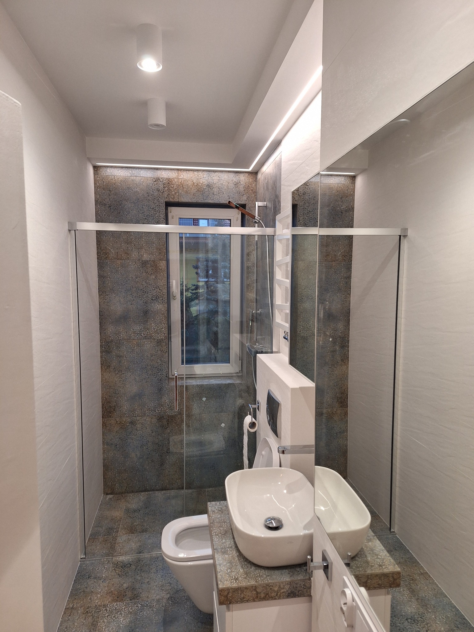 Nowoczesna łazienka z kabiną prysznicową typu walk-in, umywalką nablatową i płytkami w odcieniach szarości i brązu, oświetlona listwą LED pod sufitem.