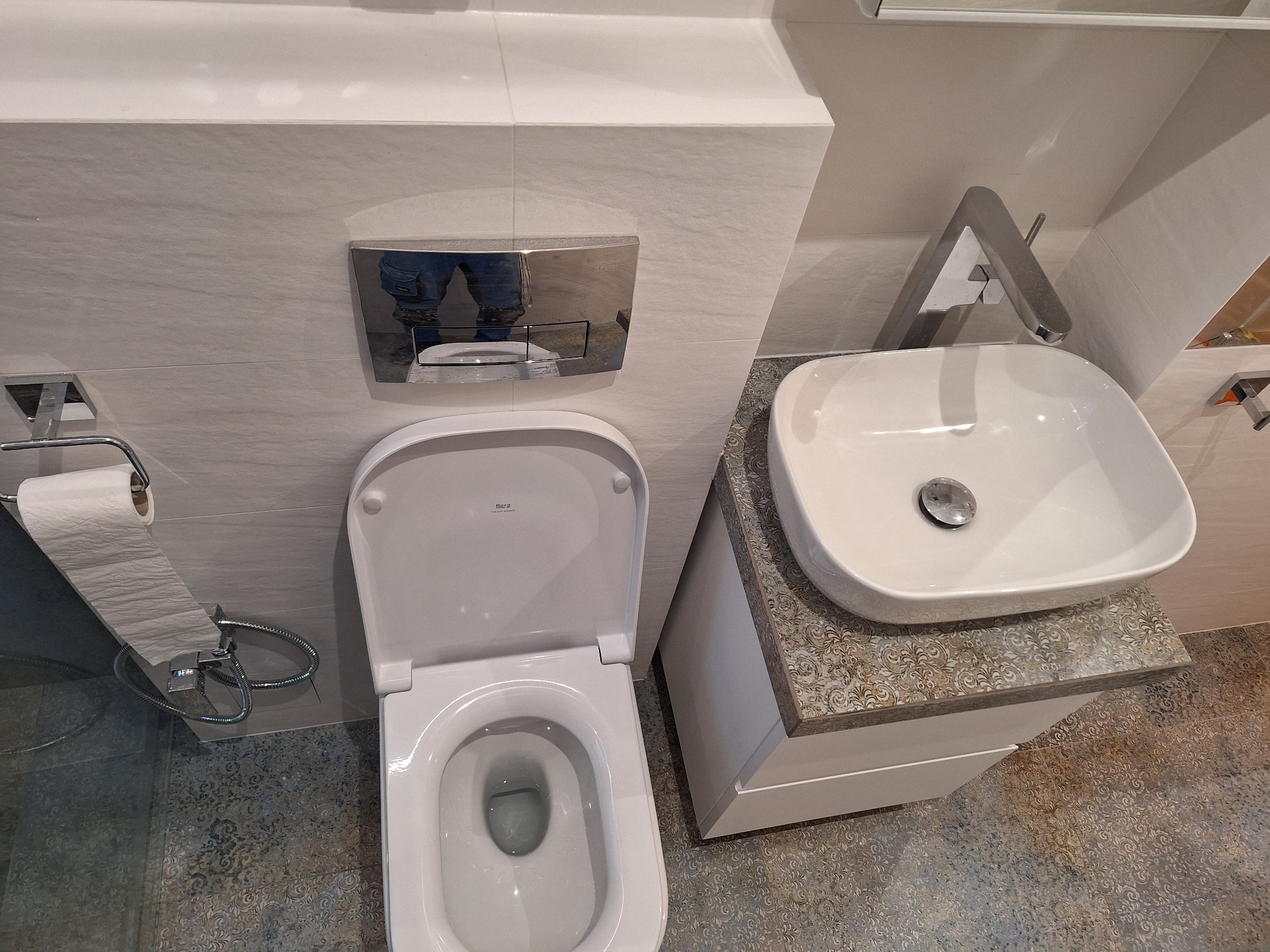 Nowoczesna łazienka z białą ceramiką: wisząca miska WC Roca, umywalka nablatowa na szafce z wzorzystym blatem i chromowana bateria, wykończona płytkami w odcieniach szarości.