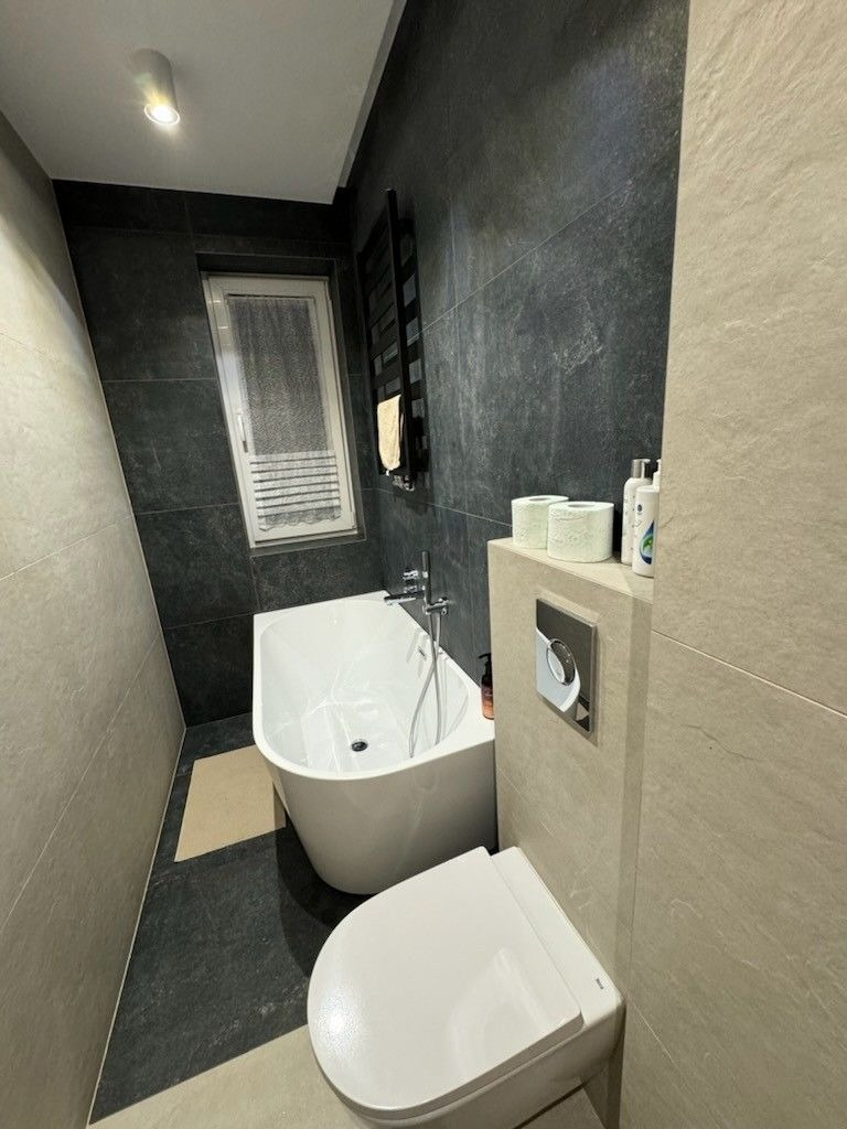 Nowoczesna łazienka z wolnostojącą wanną, podwieszaną toaletą i grzejnikiem drabinkowym na tle ciemnych, grafitowych płytek, kontrastujących z jasnymi płytkami imitującymi trawertyn.