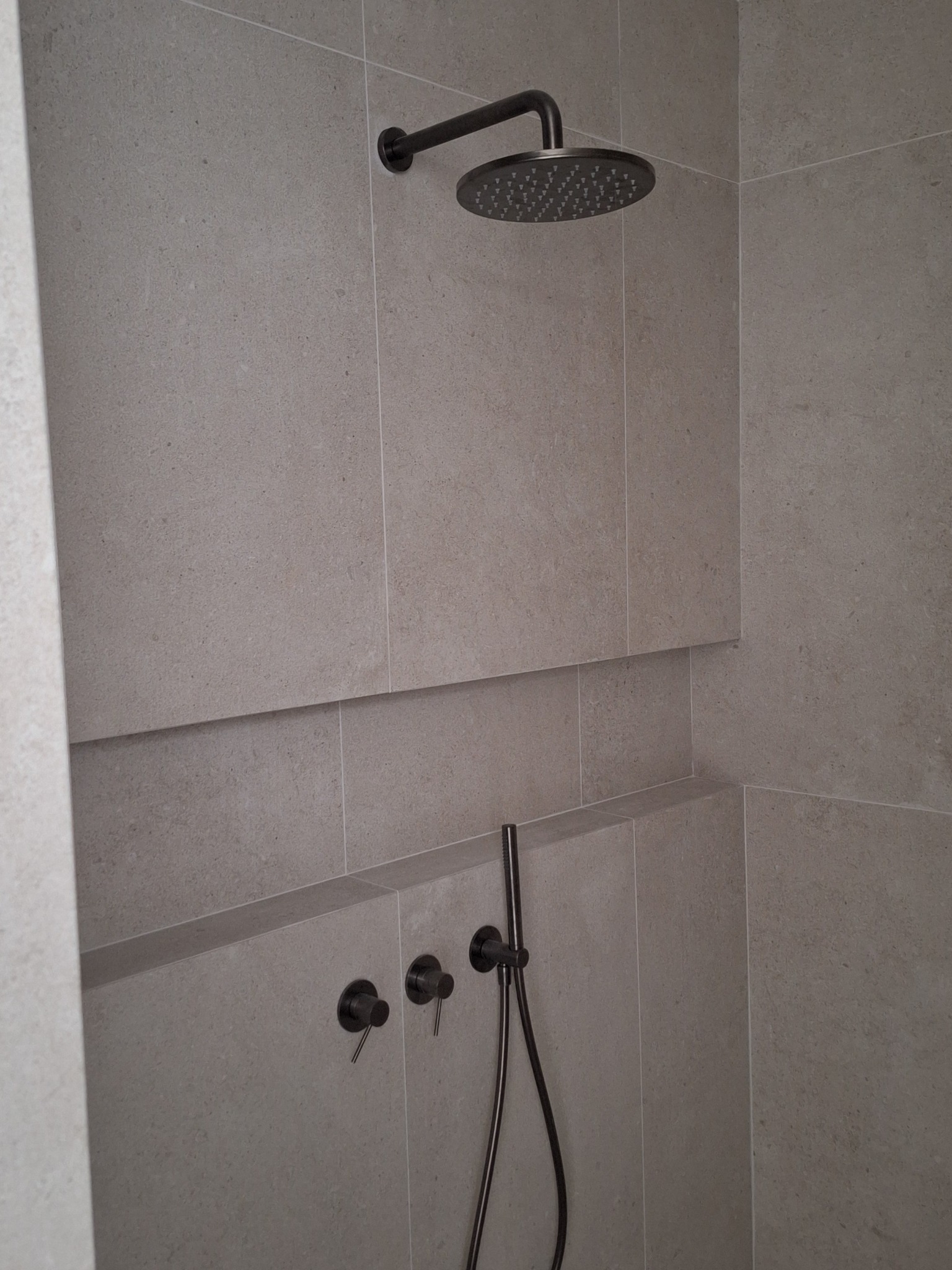 Nowoczesna kabina prysznicowa z szarymi, matowymi płytkami, deszczownicą i czarną armaturą, wnęka wykończona płytkami.