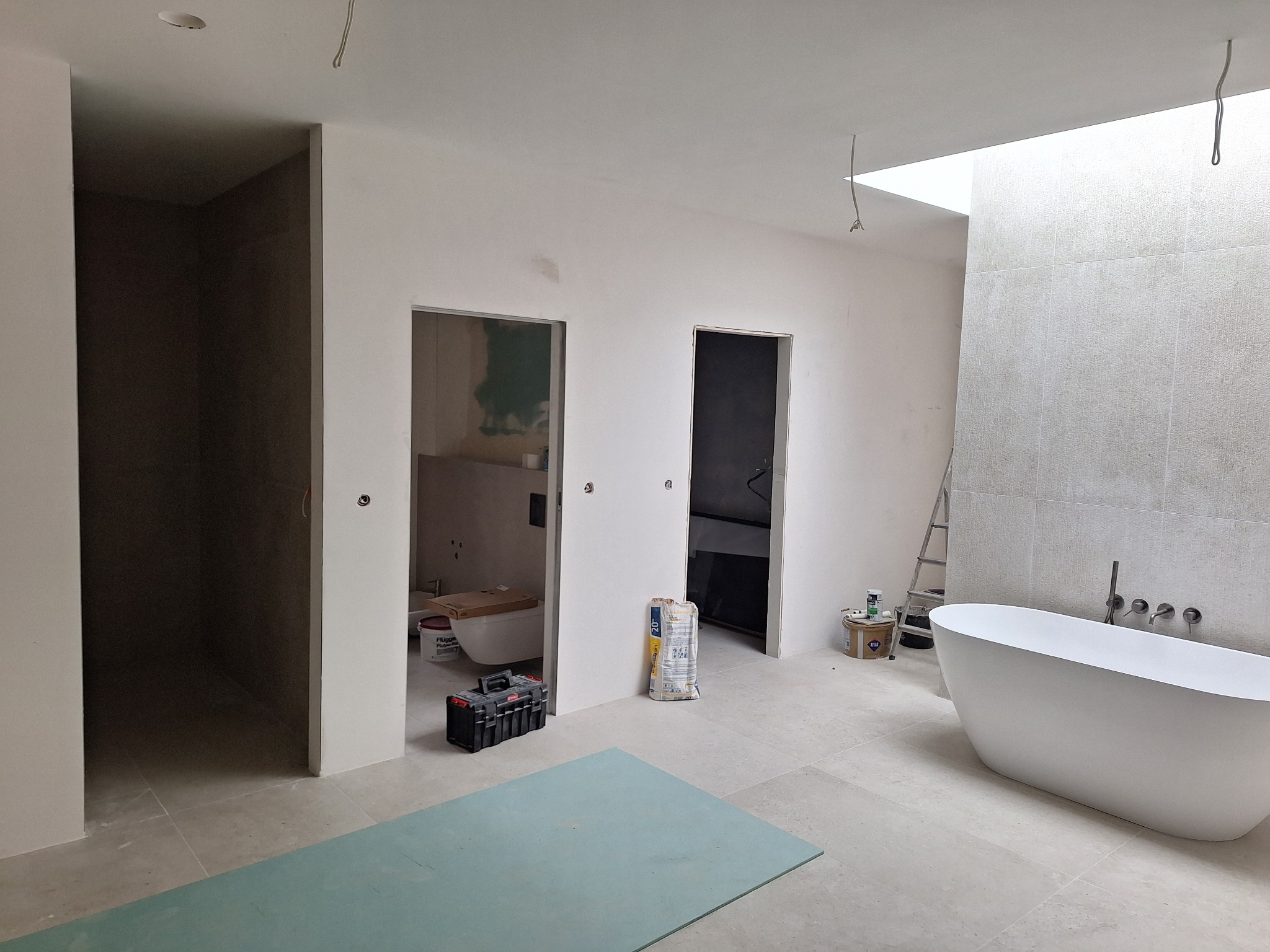 Przestronna łazienka w trakcie prac wykończeniowych, z wolnostojącą wanną, płytkami na ścianie, otworami drzwiowymi bez ościeżnic, narzędziami i materiałami budowlanymi widocznymi w tle.