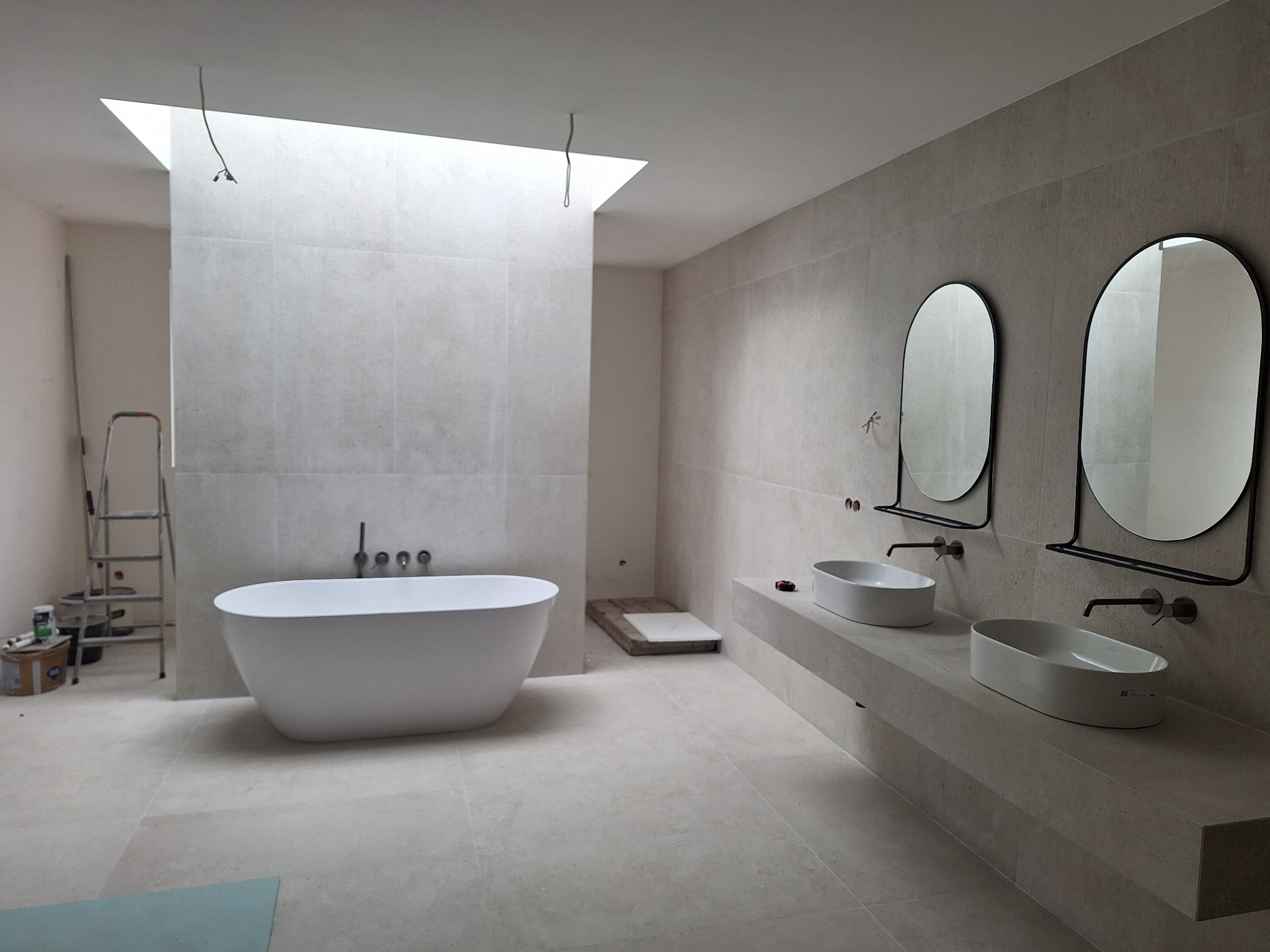 Nowoczesna łazienka w trakcie wykończenia, z wolnostojącą wanną, dwoma umywalkami nablatowymi z czarnymi bateriami i owalnymi lustrami w czarnych ramach, jasne płytki na ścianach i podłodze, surowe...