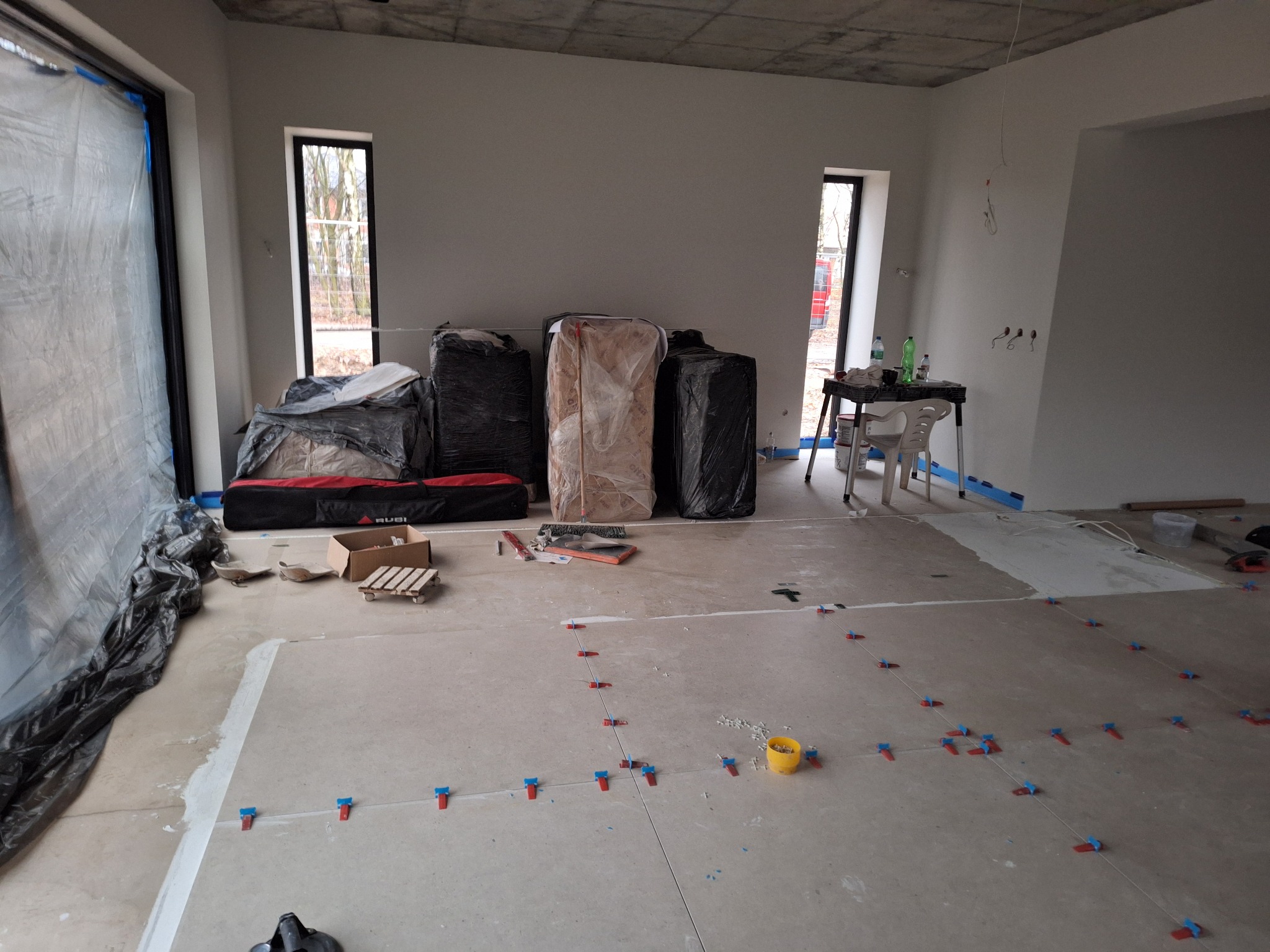 Wnętrze w trakcie układania płytek podłogowych, widoczne narzędzia glazurnicze, krzyżyki dystansowe, paczki materiałów budowlanych zabezpieczone folią oraz okna z widokiem na zewnątrz.