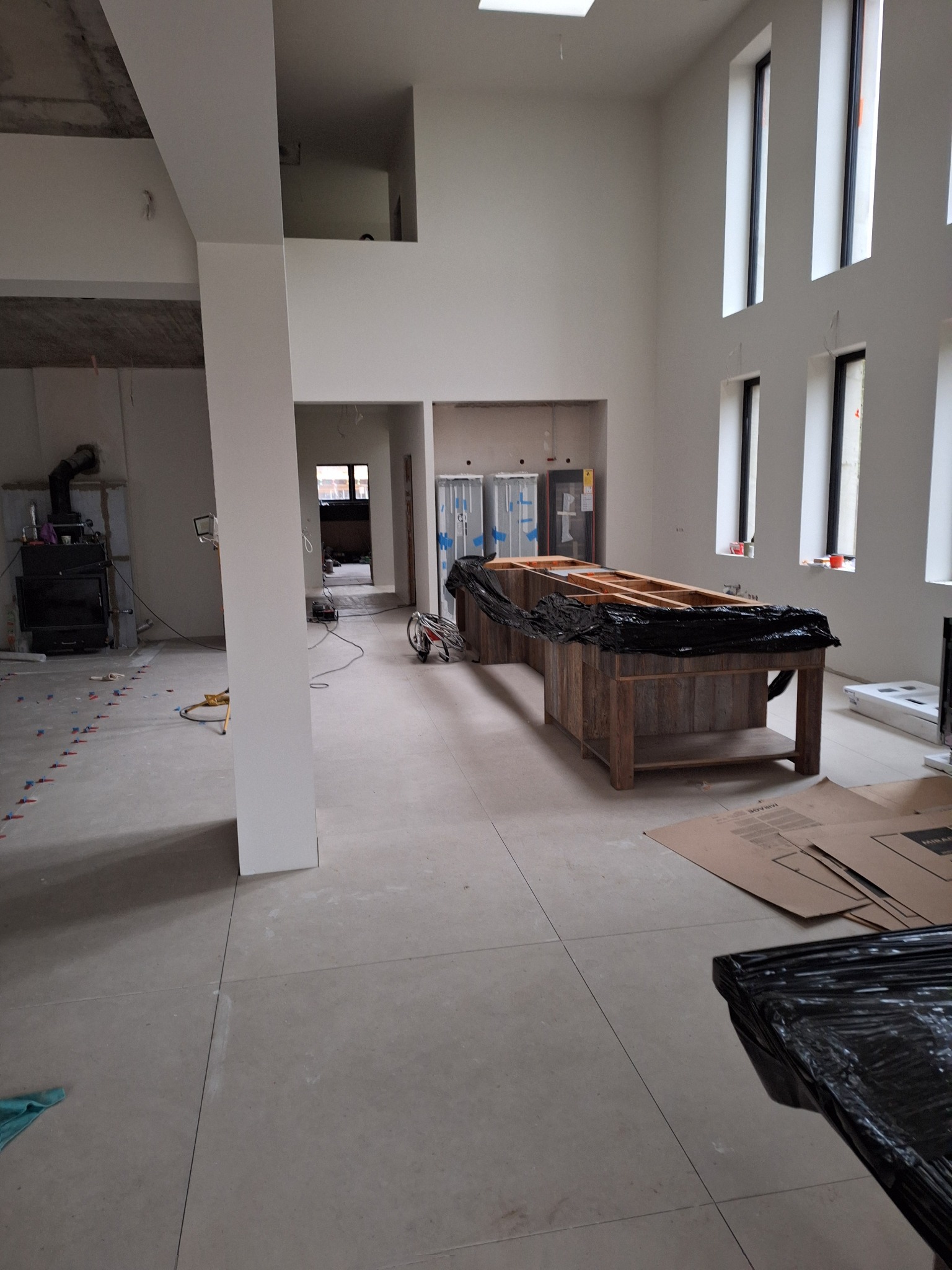 Przestronne wnętrze w trakcie prac wykończeniowych, widoczne przygotowane podłoże pod posadzkę, zabezpieczona konstrukcja wyspy kuchennej oraz wysokie okna.