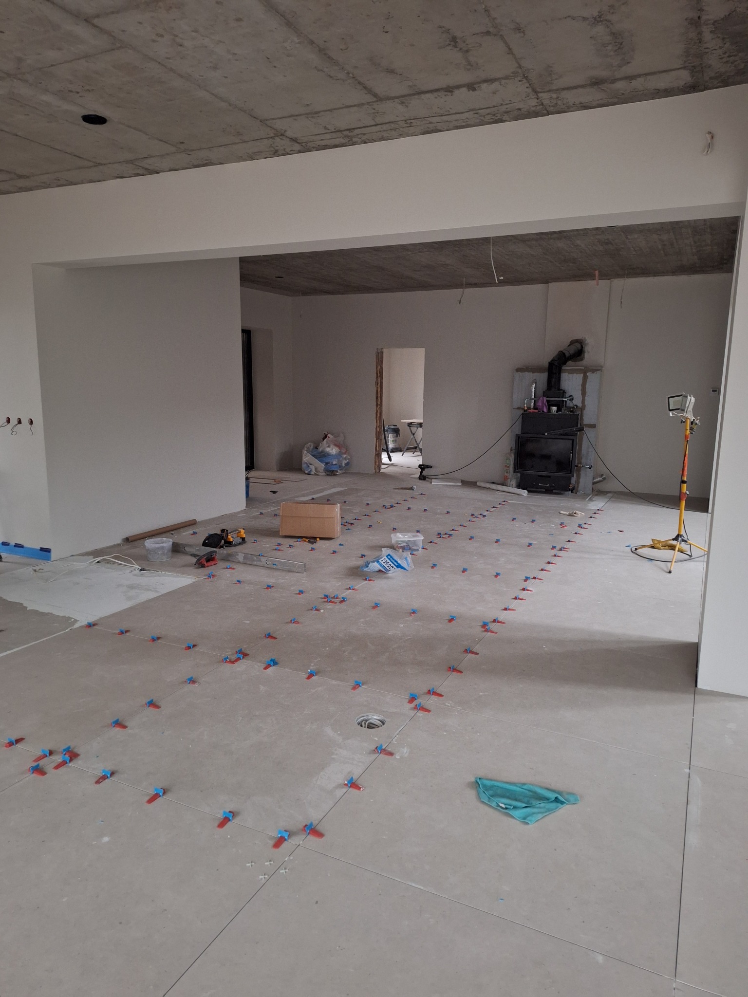 Przygotowanie do układania płytek podłogowych: rozłożone płytki z użyciem klipsów poziomujących, widoczne narzędzia i lampa budowlana w surowym wnętrzu z betonowym sufitem.