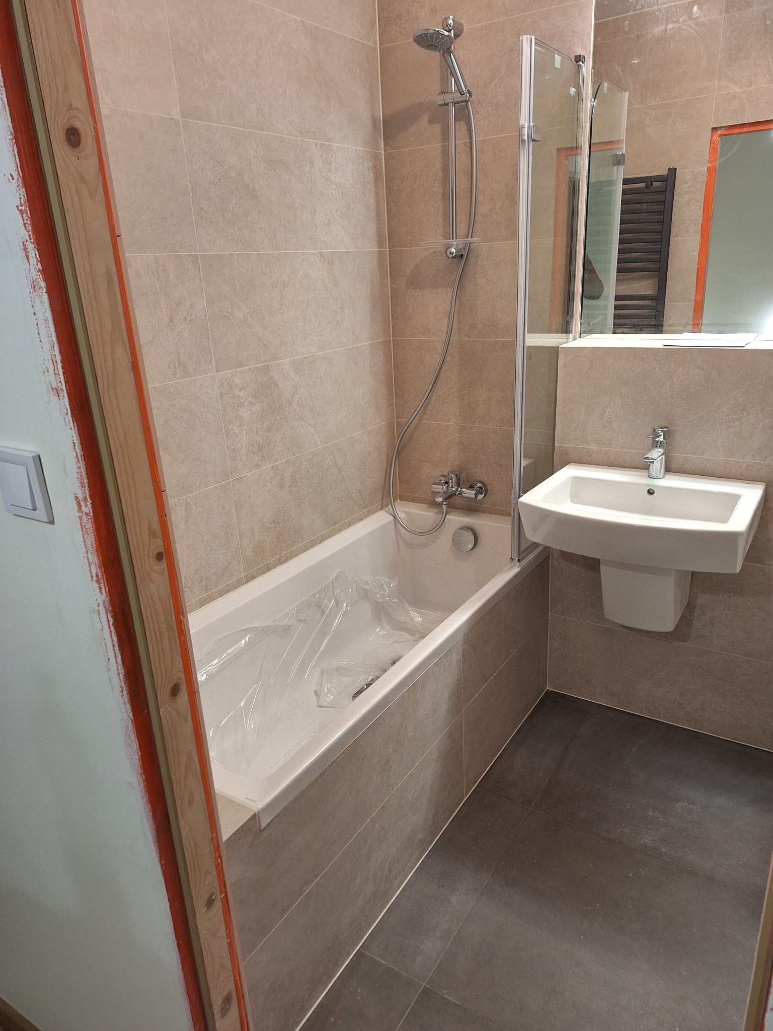 Łazienka w trakcie wykańczania: beżowe płytki ścienne, ciemna podłoga, wanna z folią ochronną, umywalka na postumencie, kabina prysznicowa ze szklaną ścianką, widoczne elementy konstrukcyjne drzwi.