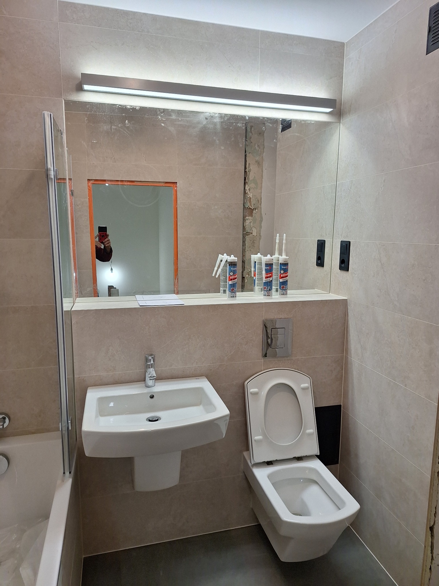 Wnętrze łazienki w trakcie prac wykończeniowych: widoczne płytki ceramiczne, umywalka, toaleta podwieszana, lustro z oświetleniem LED i częściowo odsłonięta ściana z resztkami starej farby.