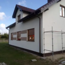 Świeżo otynkowany, dwukondygnacyjny dom z brązowymi ramami okiennymi i roletami, z widocznymi rusztowaniami i fragmentem trawnika.