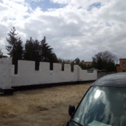 Wznoszone ściany z białych bloczków na fundamencie, widoczne zza zaparkowanego samochodu na placu budowy.