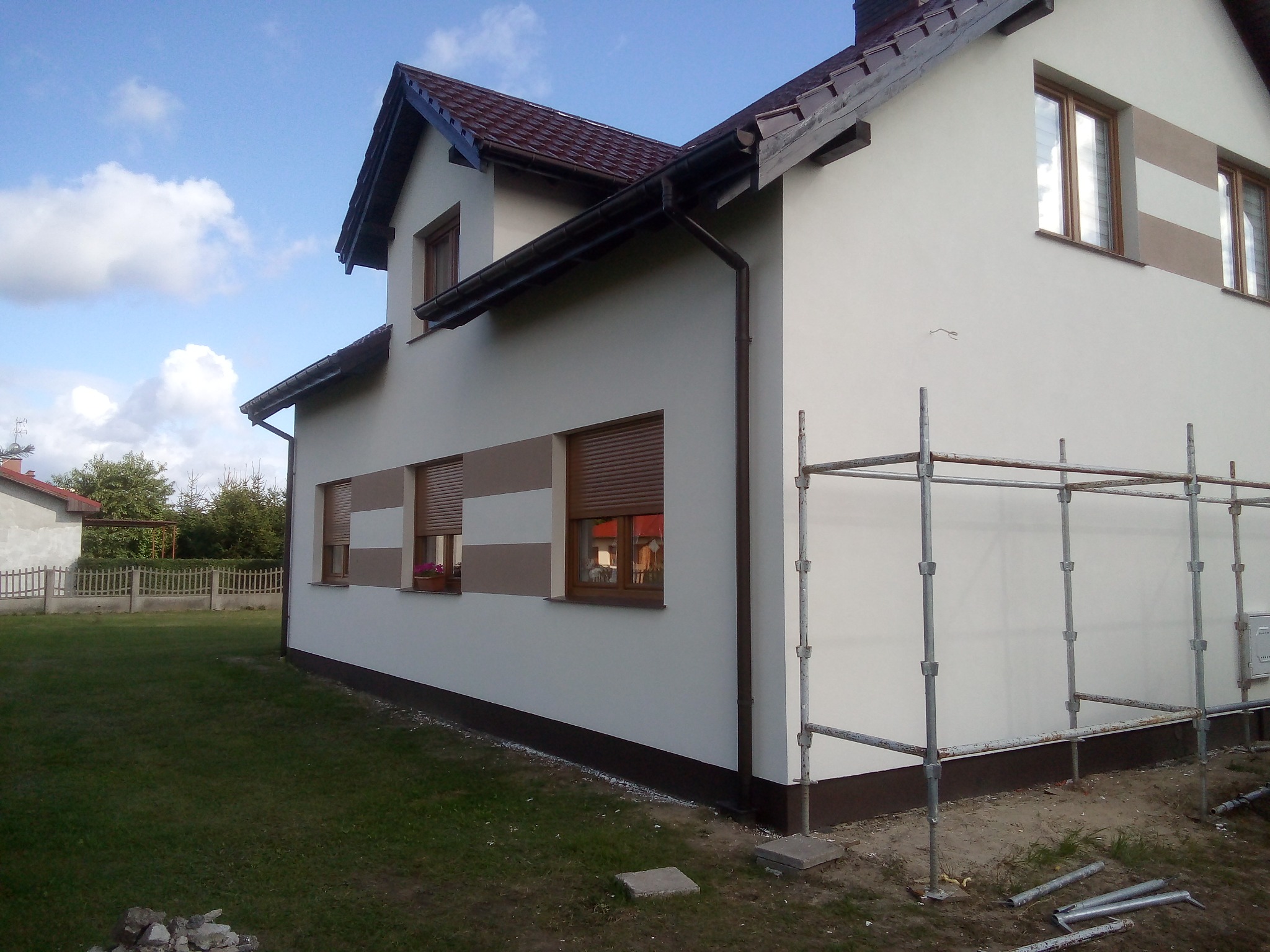 Świeżo otynkowany, dwukondygnacyjny dom z brązowymi ramami okiennymi i roletami, z widocznymi rusztowaniami i fragmentem trawnika.