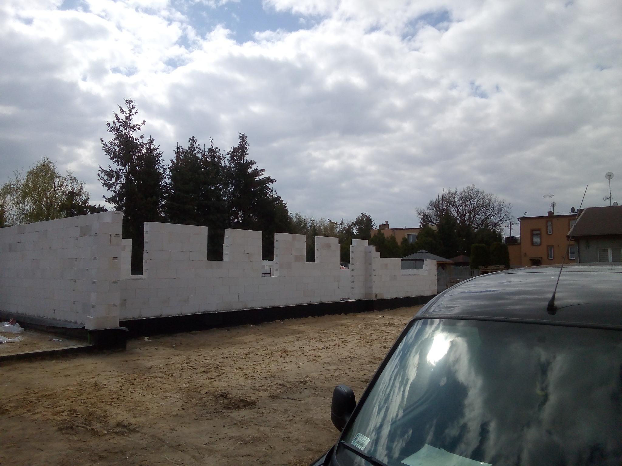 Wznoszone ściany z białych bloczków na fundamencie, widoczne zza zaparkowanego samochodu na placu budowy.