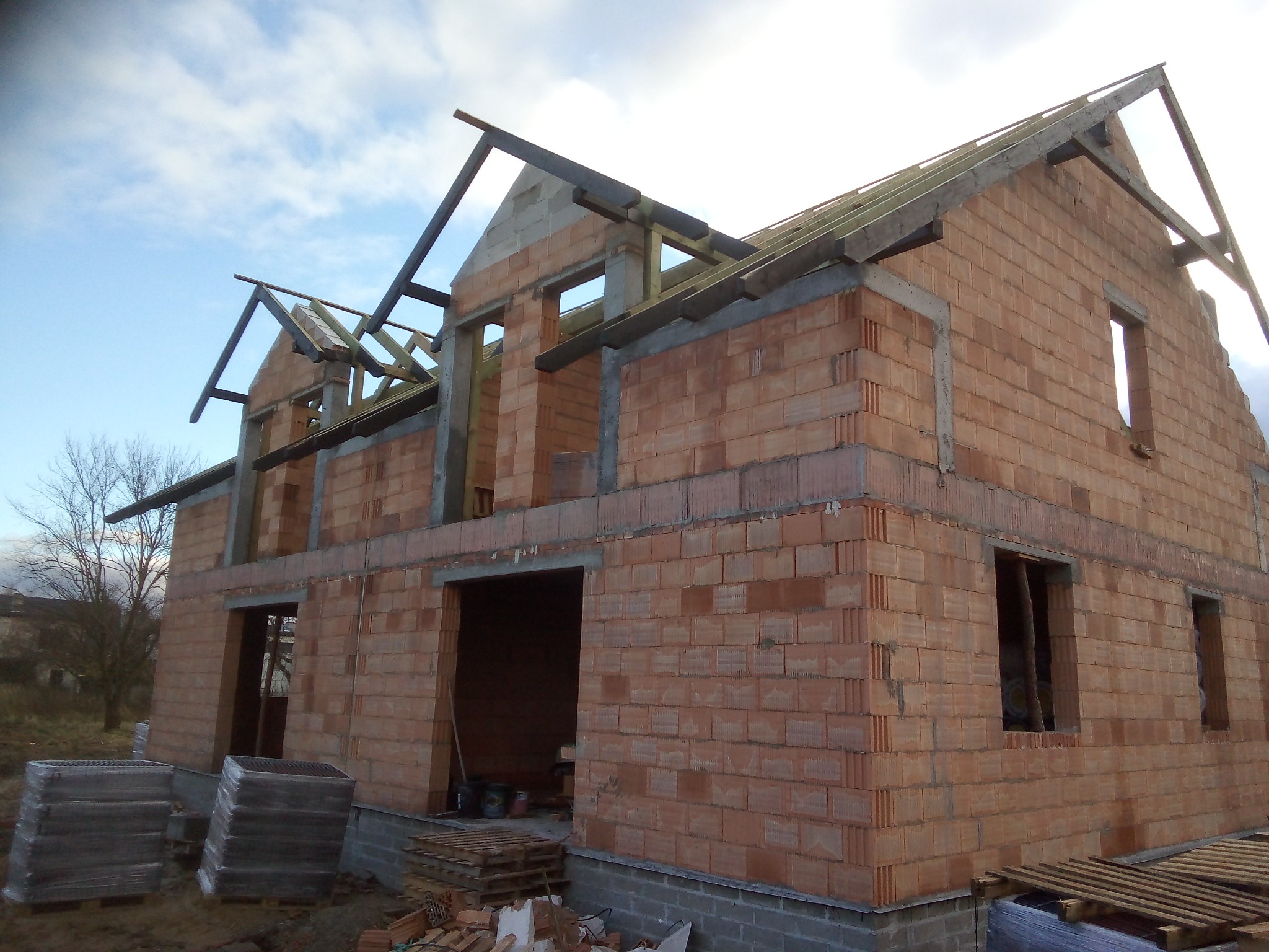 Surowy stan domu murowanego z czerwonej cegły, widoczne niezadaszone ściany z otworami okiennymi i drzwiowymi, drewniana konstrukcja dachu w trakcie montażu, palety z materiałami budowlanymi przed...