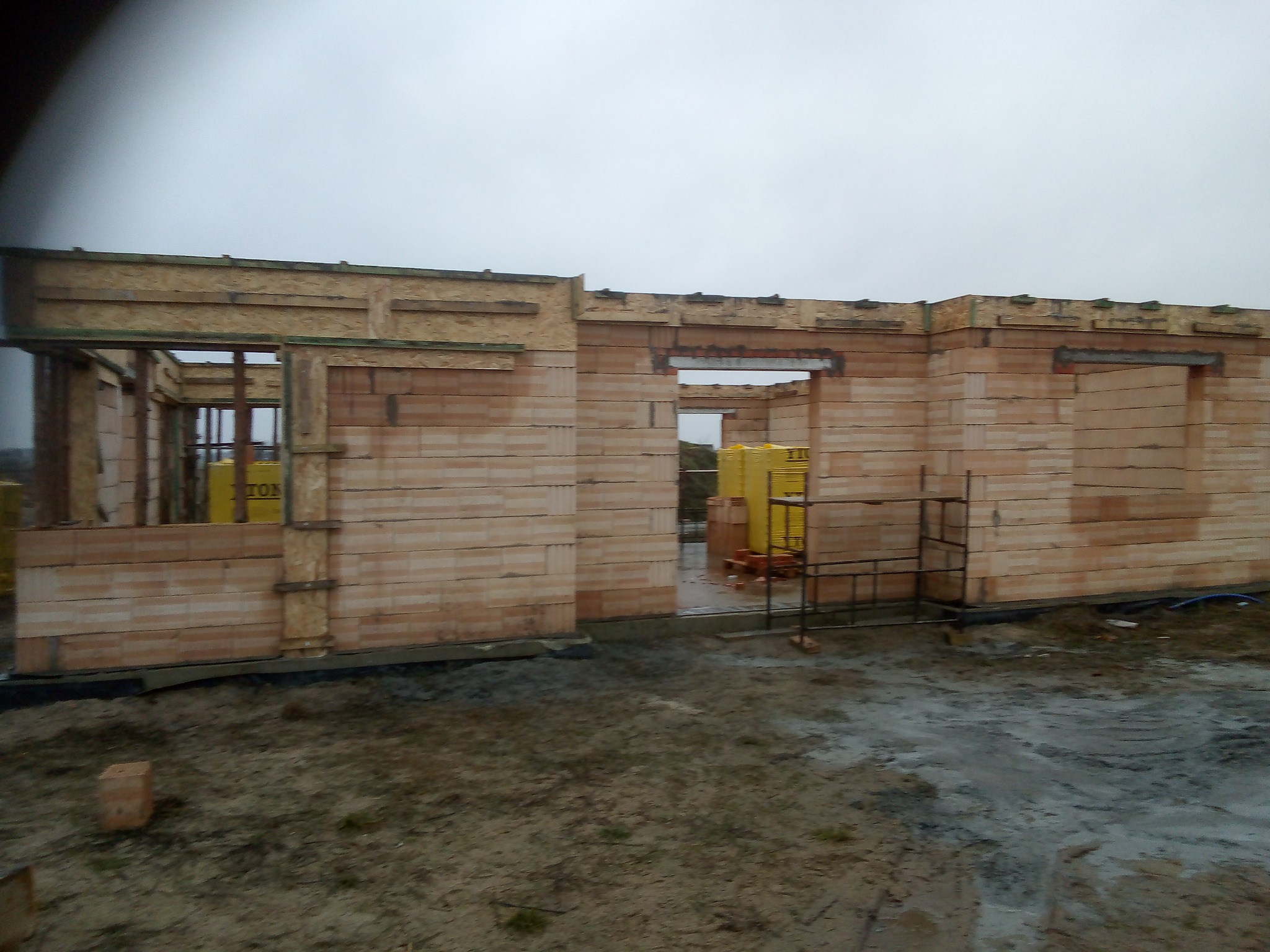 Wznoszenie ścian parterowego domu z bloczków ceramicznych, widoczne otwory okienne i drzwiowe, paleta z bloczkami w tle, teren budowy w trakcie prac.