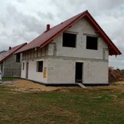 Surowy, dwukondygnacyjny dom w trakcie budowy z czerwonym dachem, widoczne białe bloczki konstrukcyjne, nieotynkowane ściany, przygotowane otwory okienne i drzwiowe, obok sterta materiałów...