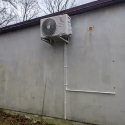 Zewnętrzna jednostka klimatyzacji Rotenso Mirai z WiFi M35X R15 zamontowana na szarej ścianie budynku ze spadzistym dachem, widoczne rury odprowadzające skropliny.