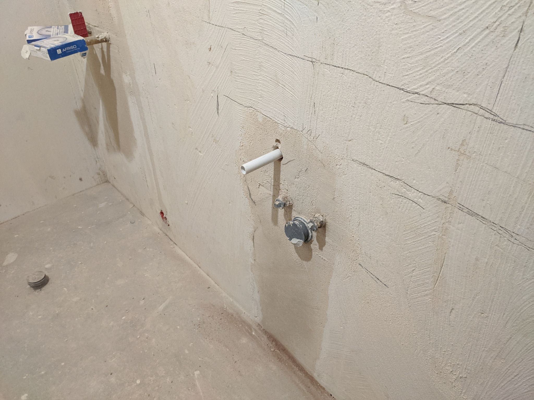 Surowa ściana w stanie deweloperskim z widocznymi rurami instalacji wodnej i zaworami, przygotowana do dalszych prac wykończeniowych, zarysowane linie na ścianie, pudełko z elementami montażowymi...
