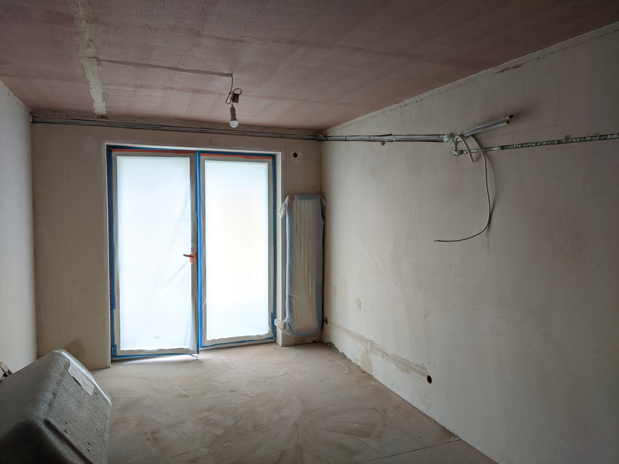 Wnętrze mieszkania w stanie deweloperskim z widoczną instalacją klimatyzacji ukrytą w ścianach i odprowadzeniem skroplin do kanalizacji, rury prowadzone pod sufitem, okno zabezpieczone folią malarską.