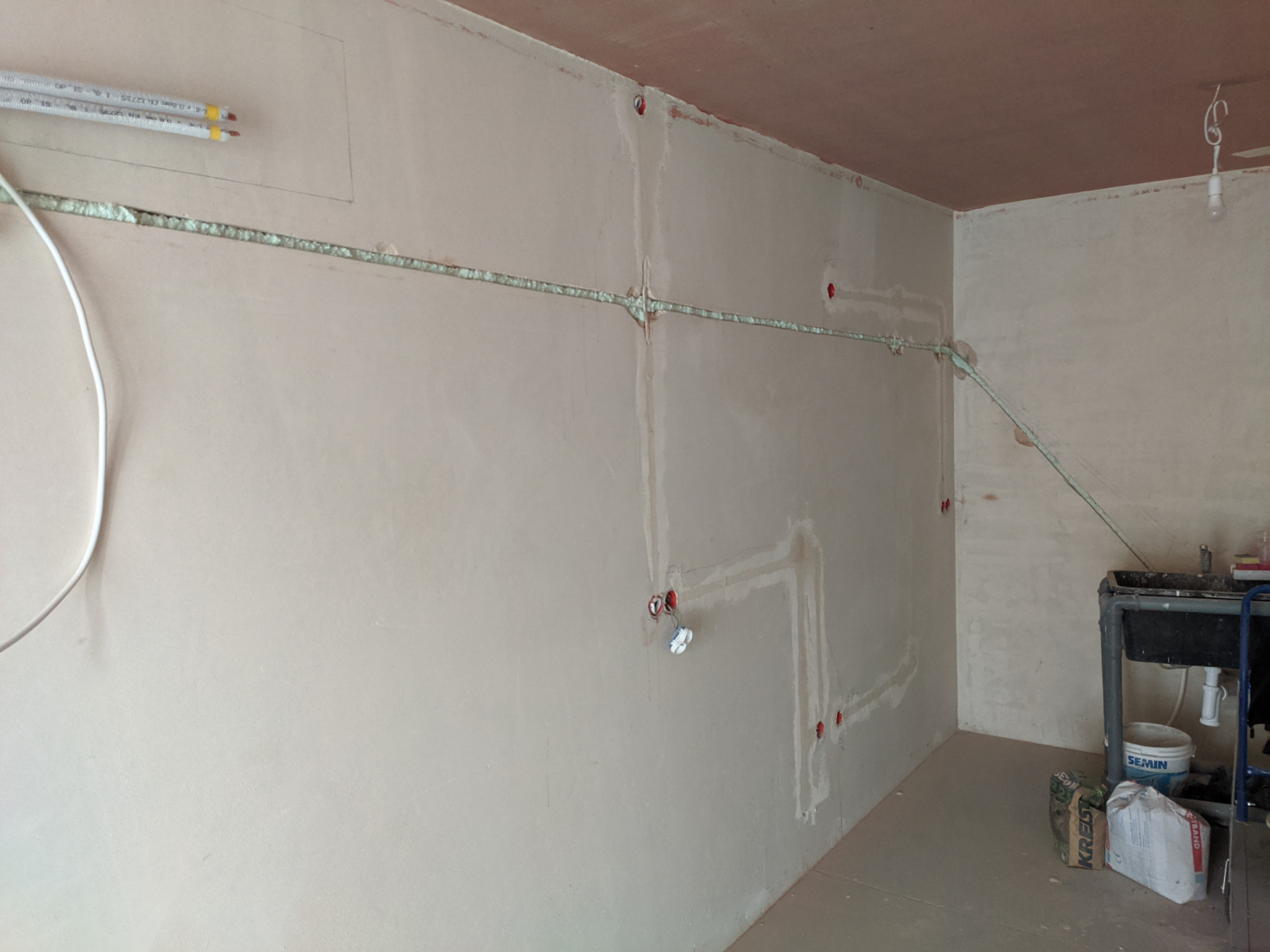 Instalacja elektryczna w ścianach mieszkania w stanie deweloperskim, widoczne bruzdy wypełnione pianką montażową, puszki elektryczne i przewody. W rogu pomieszczenia prowizoryczny zlew i worki...