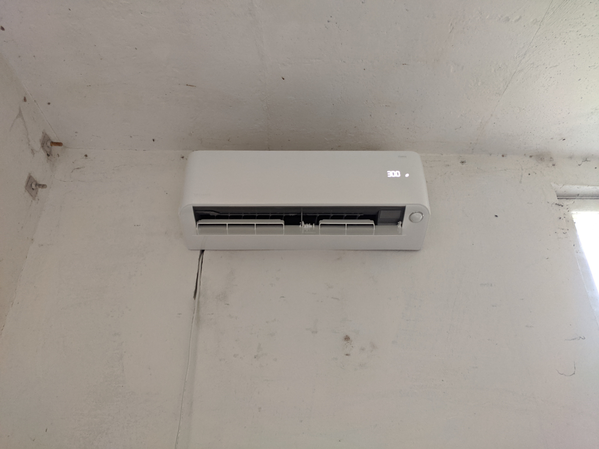 Biały klimatyzator ścienny zamontowany na pomalowanej na biało ścianie z widocznymi śladami po instalacji i pęknięciami w tynku.