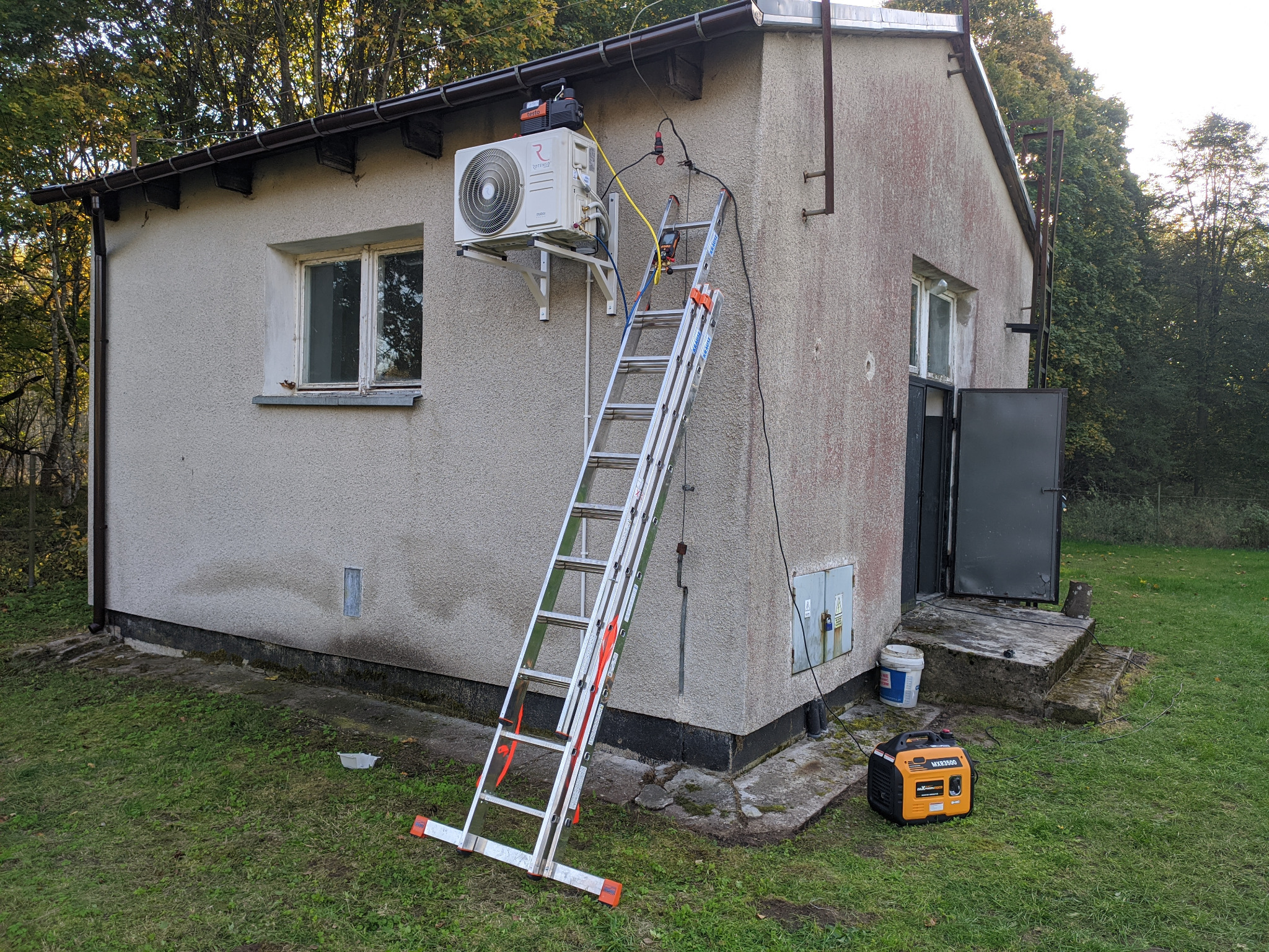 Zewnętrzna jednostka klimatyzacji zamontowana na ścianie budynku, podłączona do agregatu prądotwórczego inwerterowego stojącego na trawie, z użyciem drabiny rozstawionej na ziemi.