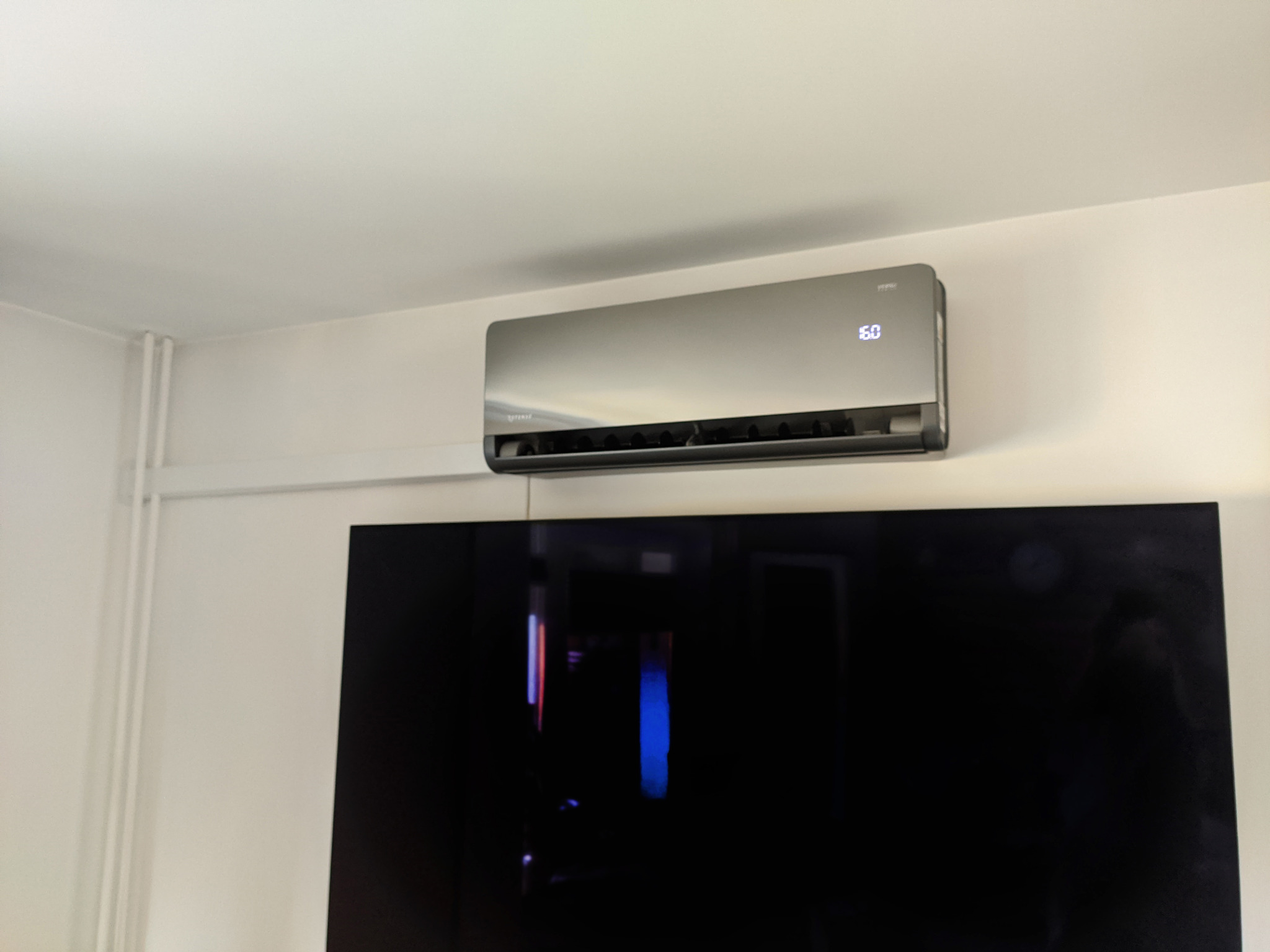 Zamontowana srebrna klimatyzacja ścienna z wyświetlaczem temperatury 16 stopni Celsjusza, zainstalowana nad dużym czarnym telewizorem.