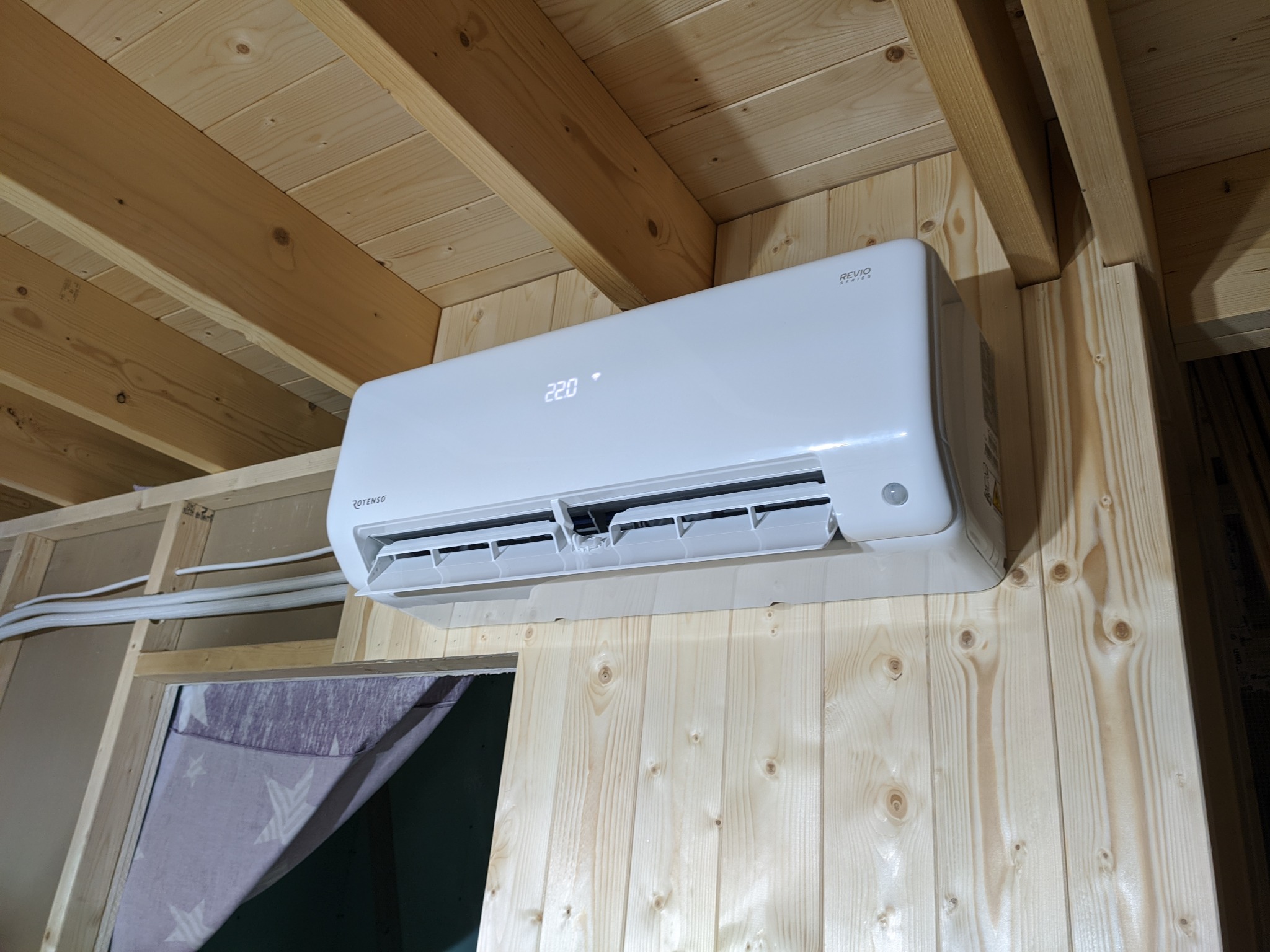 Biały klimatyzator Rotenso Revio RO35X R14 zamontowany na drewnianej ścianie w domku letniskowym, widoczny wyświetlacz temperatury 22.0 stopnie Celsjusza i fragment instalacji elektrycznej.