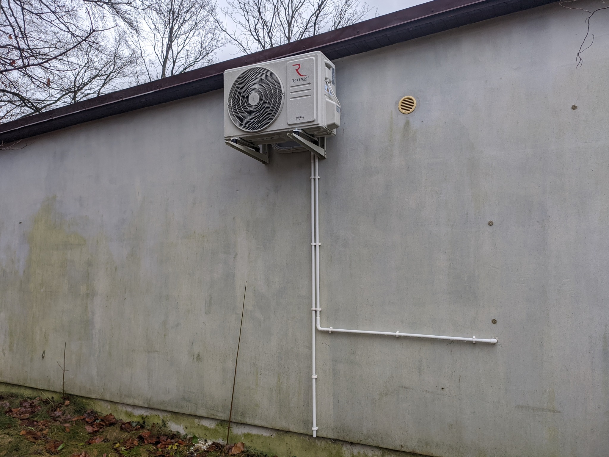 Zewnętrzna jednostka klimatyzacji Rotenso Mirai z WiFi M35X R15 zamontowana na szarej ścianie budynku ze spadzistym dachem, widoczne rury odprowadzające skropliny.