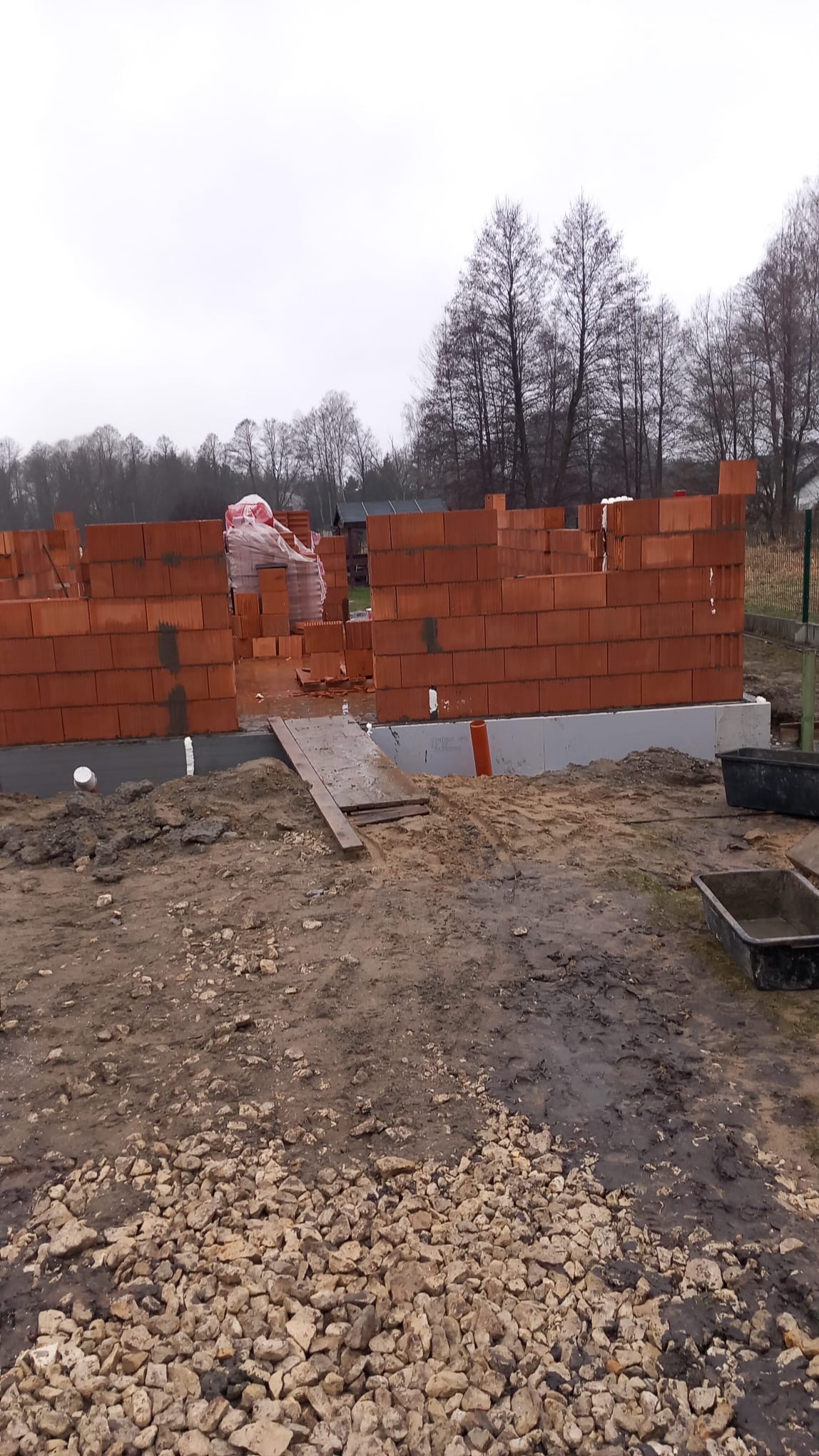 Budowa domu w trakcie: ściany z czerwonej cegły wzniesione na fundamencie, widoczne materiały budowlane i otoczenie placu budowy w pochmurny dzień.