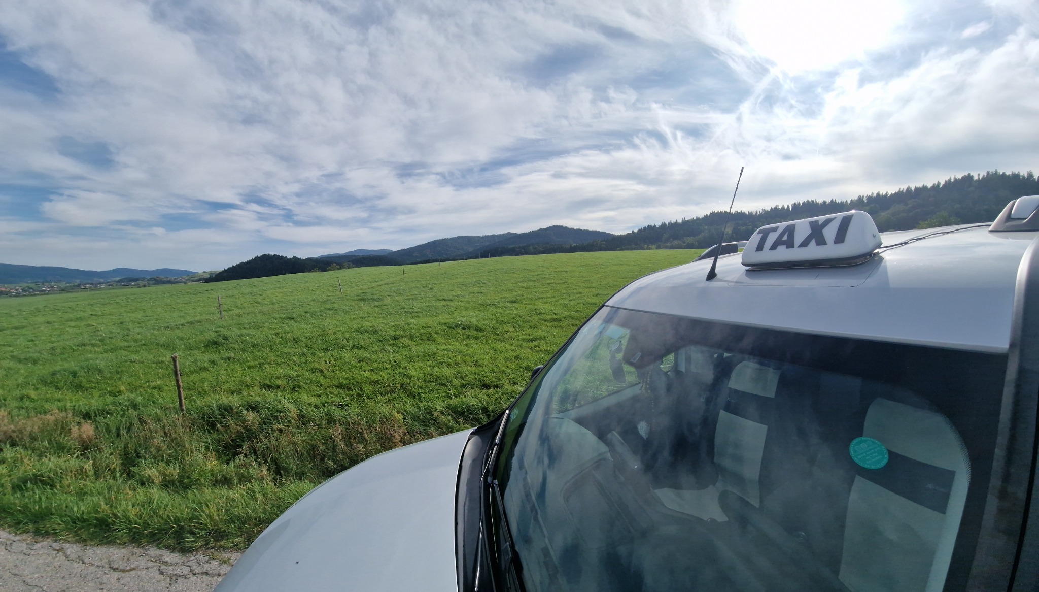 Taxi z lampą 'TAXI' na dachu, stojące na skraju drogi z widokiem na zielone wzgórza i błękitne niebo z chmurami.