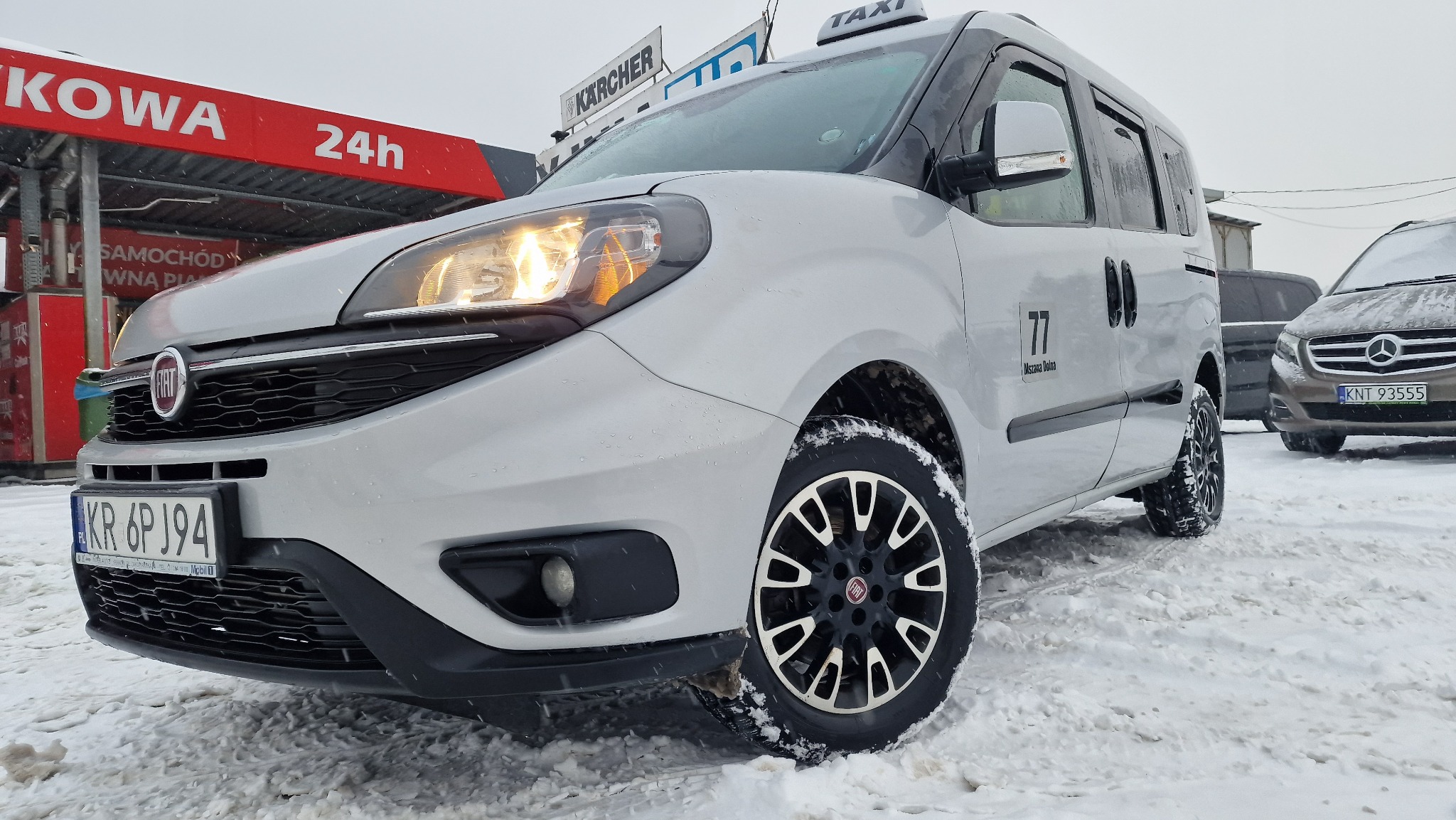 Biały Fiat Doblo Taxi na śniegu, z włączonymi światłami, zaparkowany przed stacją benzynową Kowa, widok z niskiej perspektywy.